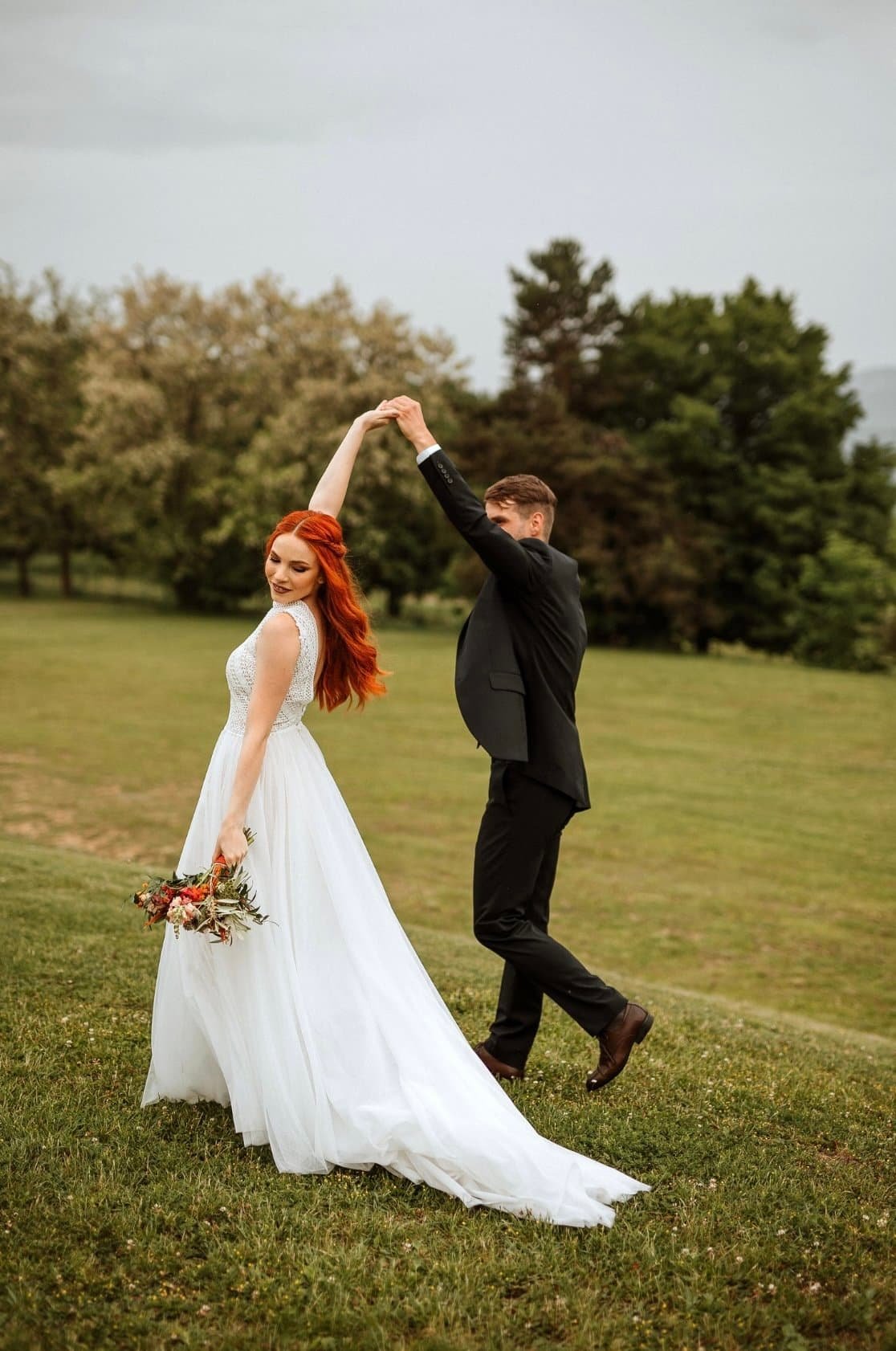 A bride and groom dancing outdoors on a grassy field, with the bride holding a bouquet and the groom wearing a black suit. The groom is twirling the bride, who is smiling, while holding her hand up.