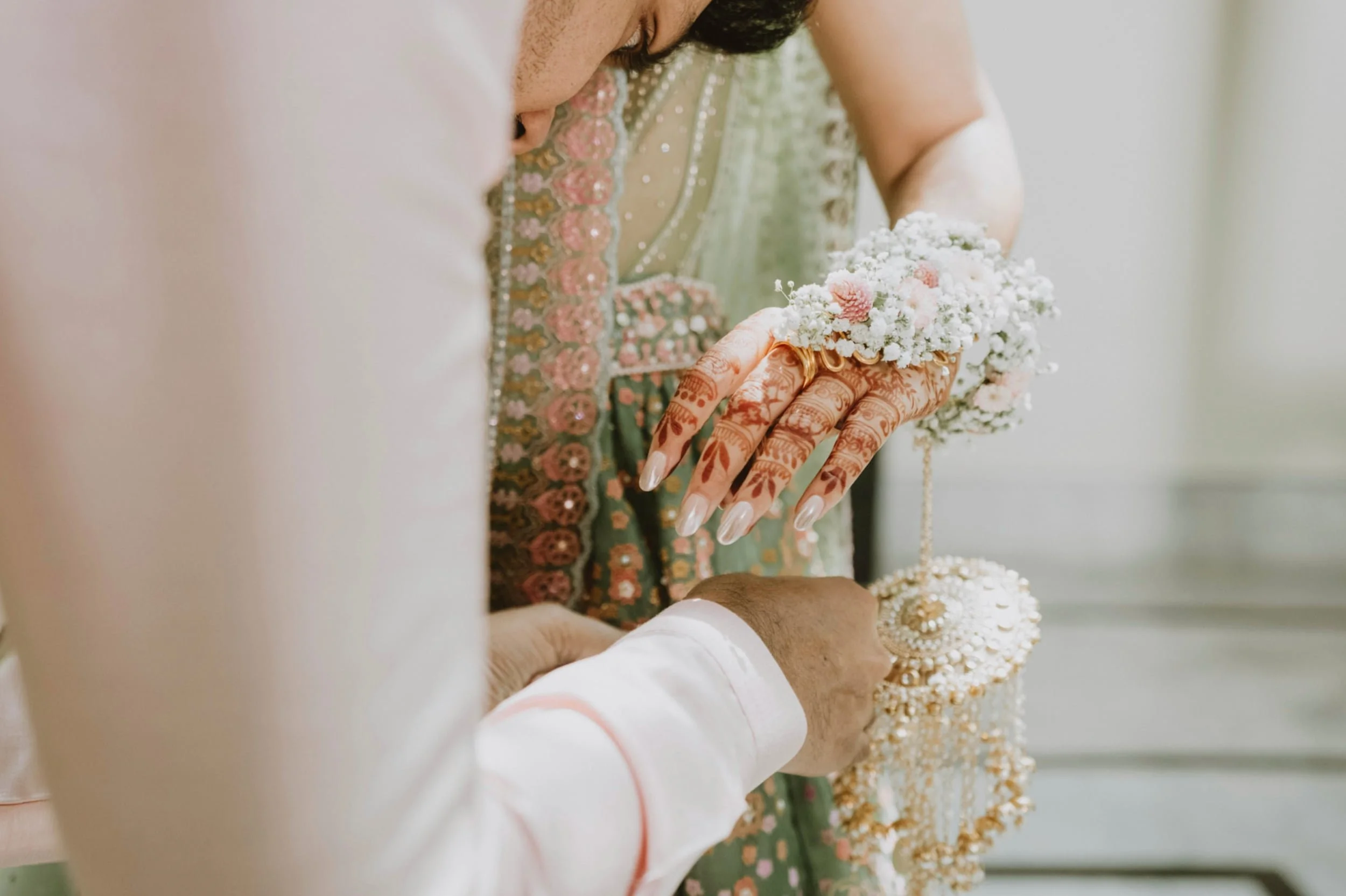 A woman dressed in traditional Indian attire, with henna on her hands, holding a small floral jewelry stand or decoration, possibly during a celebration or ceremony.