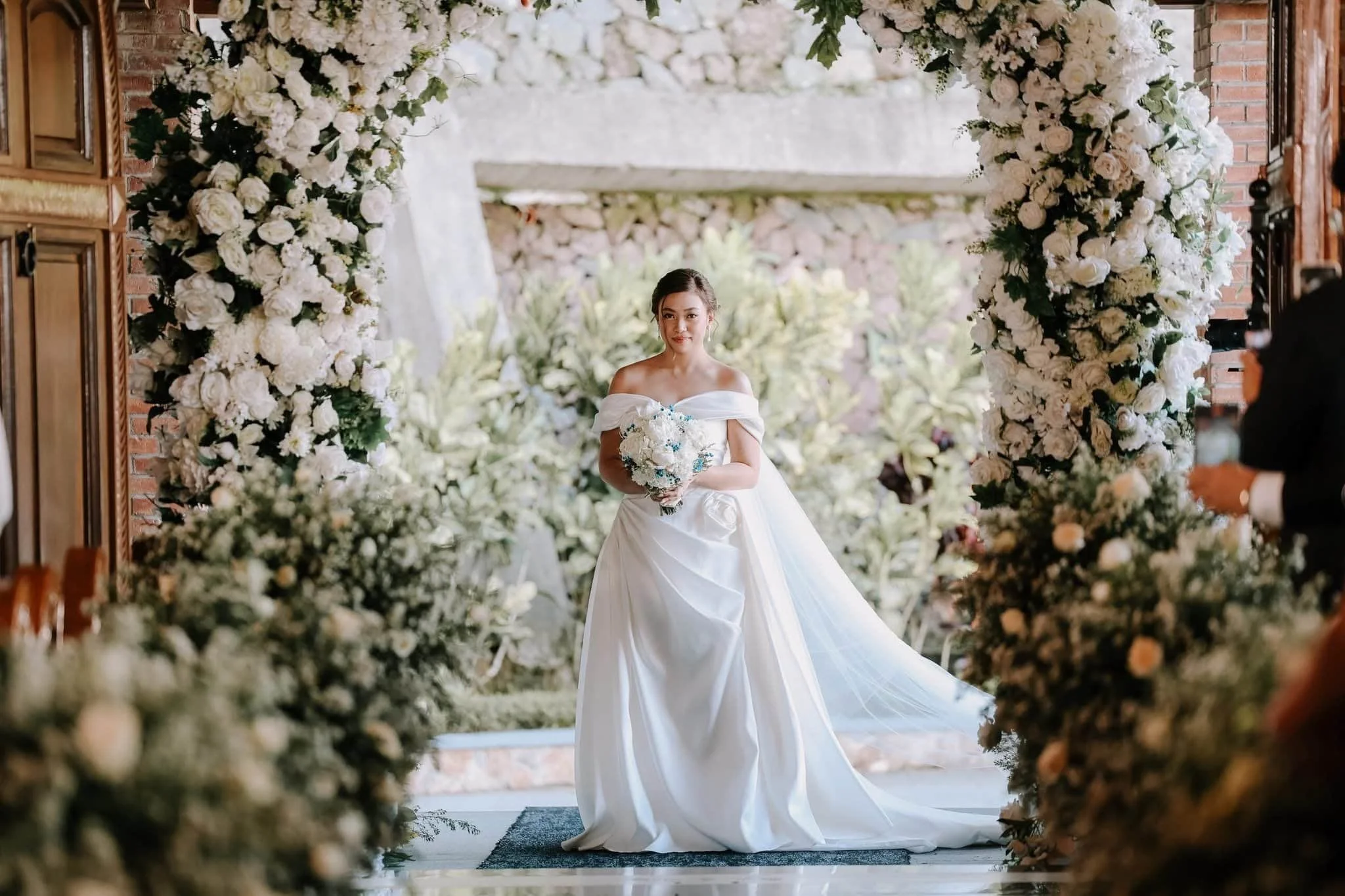 A bride in a white off-shoulder wedding gown holding a bouquet of white flowers standing under a floral arch at a wedding ceremony. The arch is decorated with white roses and greenery, with trees and a stone wall background.