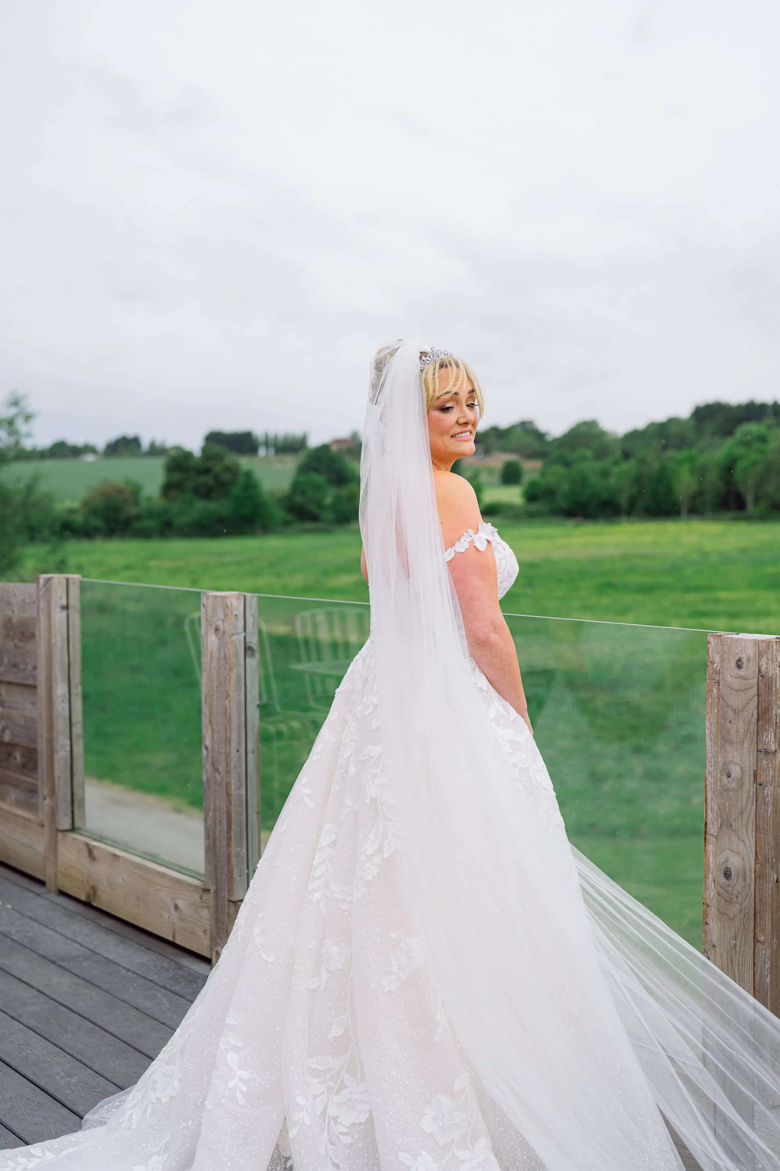 A bride in a white wedding dress with a long veil outdoors on a cloudy day, standing near a wooden railing with green fields and trees in the background.