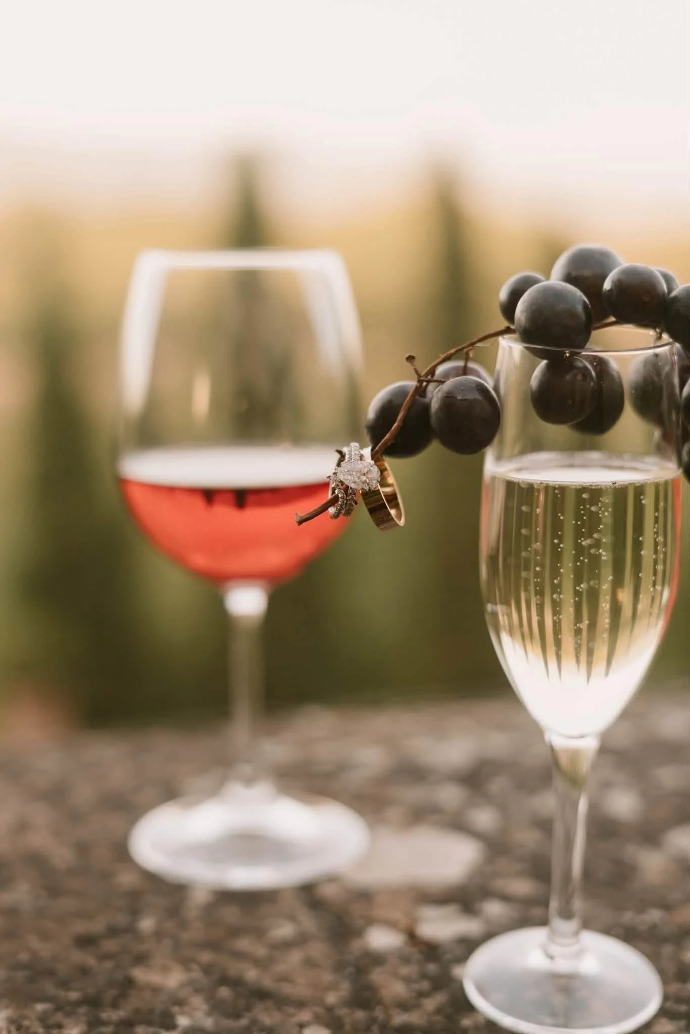 Two glasses of sparkling wine, one with a light pink color and the other with a pale golden color, are on a granite countertop. One glass is decorated with a bunch of dark grapes and an engagement ring placed on the grapes.