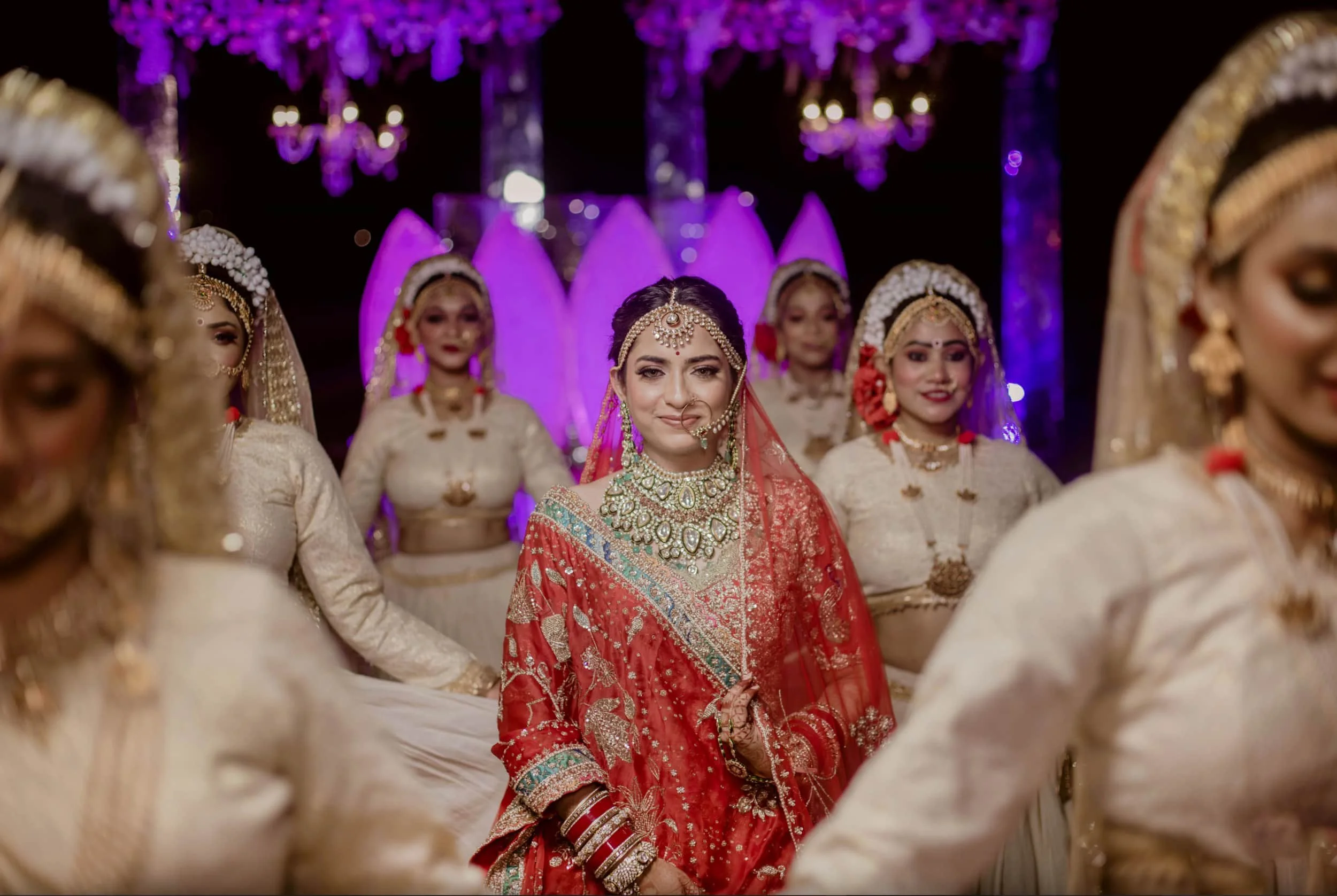 A bride in a red wedding dress with intricate gold jewelry and headpieces, surrounded by bridesmaids in cream and gold outfits, at night with purple lighting and chandeliers in the background.