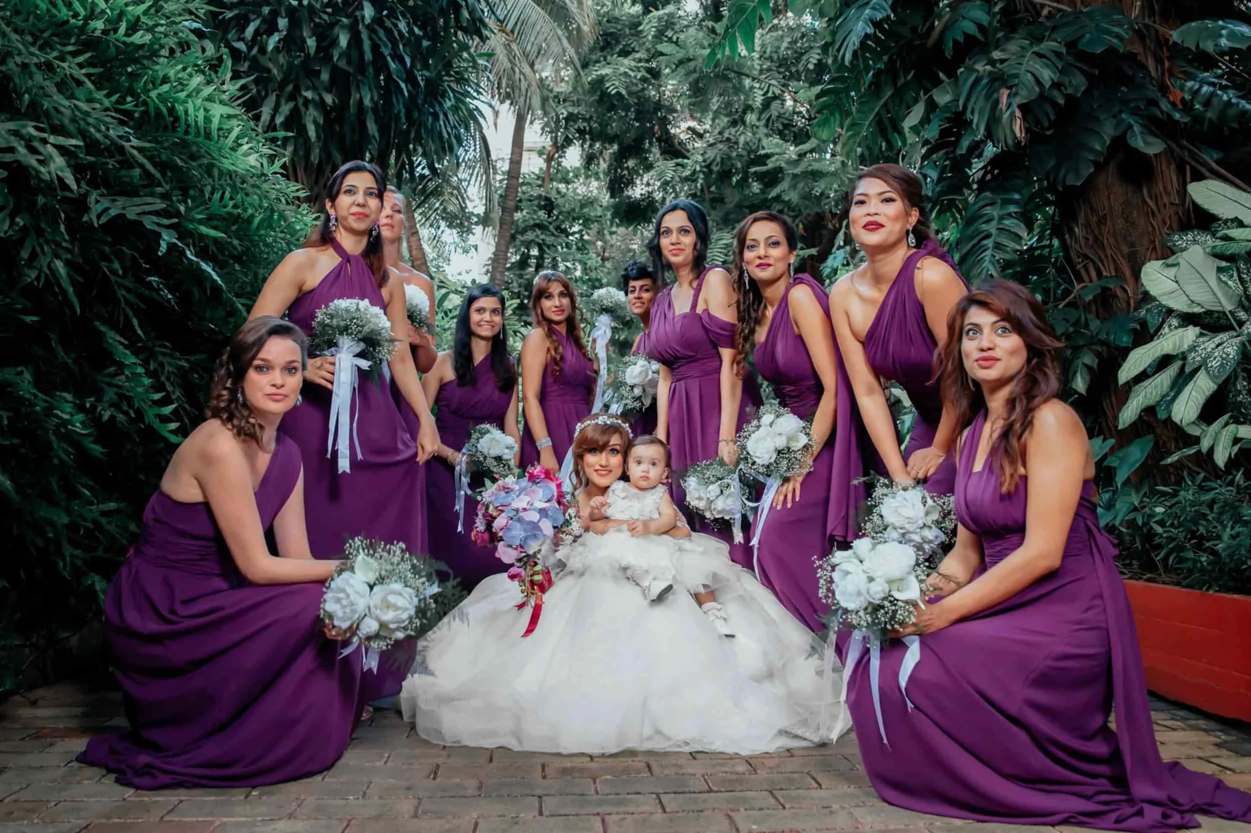 A group of women and two young girls dressed in purple gowns, gathered outdoors among lush green tropical plants, for a special occasion, possibly a wedding or celebration.