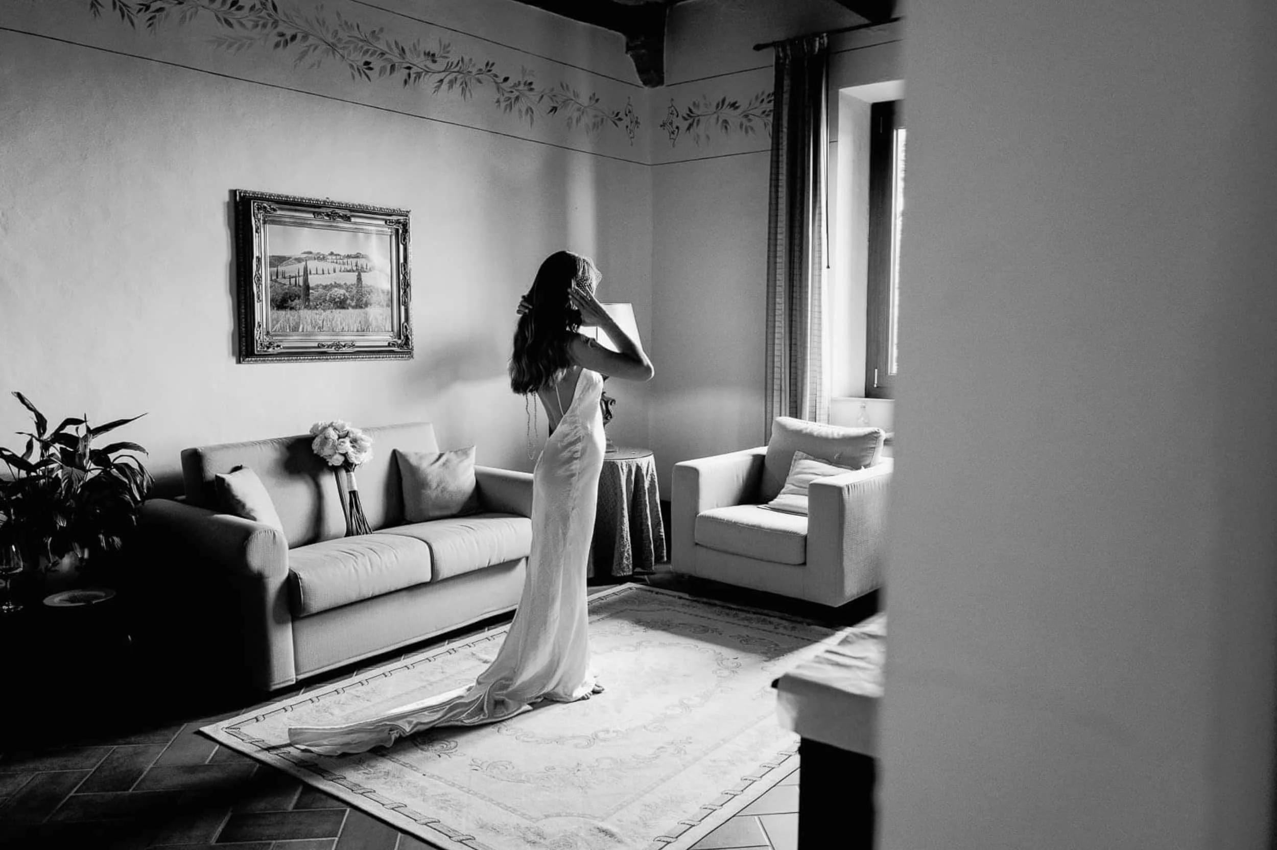 A woman in a wedding dress standing in a living room, adjusting her hair while looking at her reflection. The room has a sofa, a painting on the wall, a potted plant, and curtains by a window.