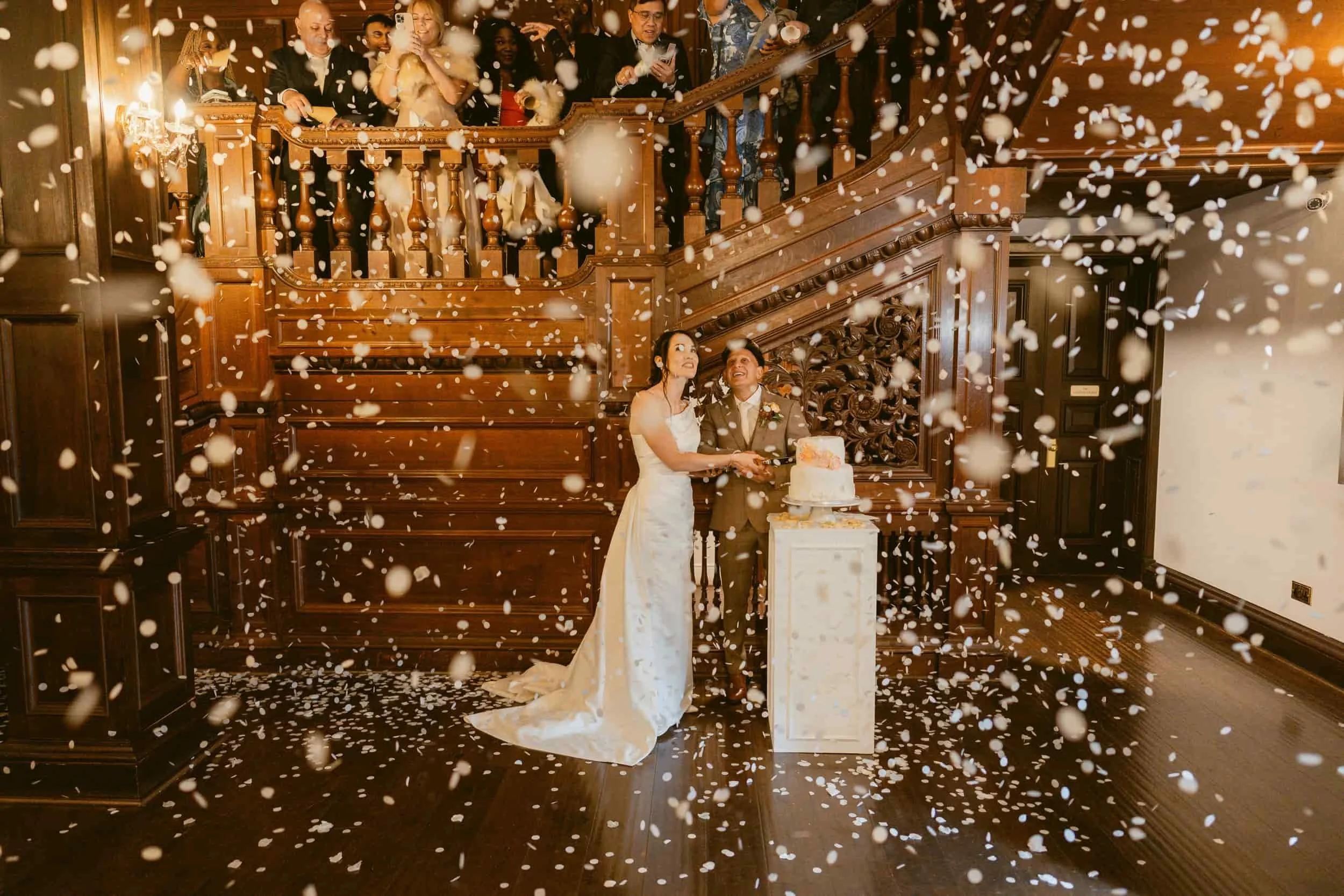Wedding reception with a bride and groom cutting their wedding cake amidst falling confetti and guests watching from the staircase.