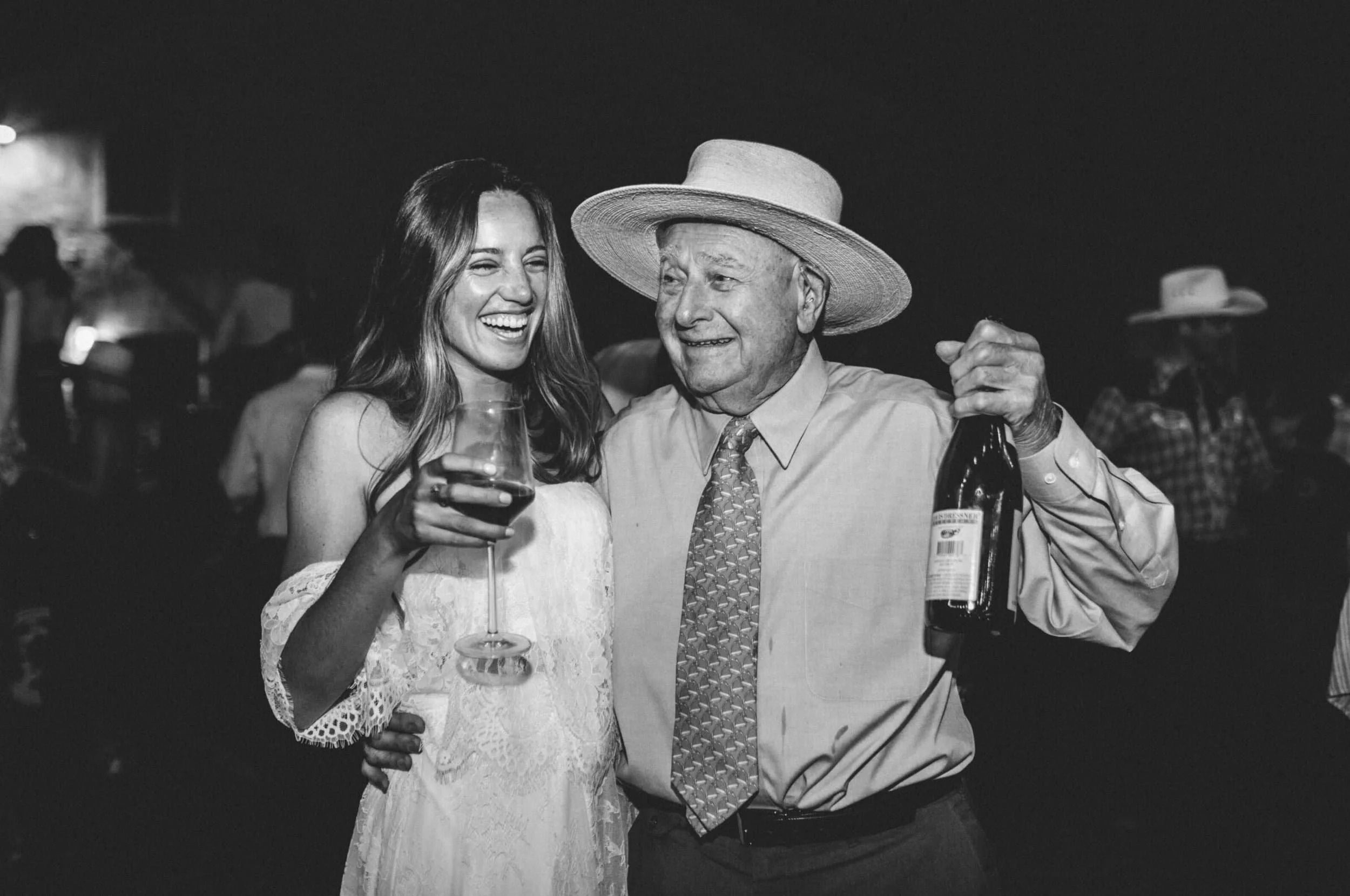 A black and white photo of a young woman and an elderly man sharing a joyful moment at a social gathering. The woman is holding a glass of wine and laughing, while the man, wearing a large straw hat and holding a bottle, is smiling warmly at her. The