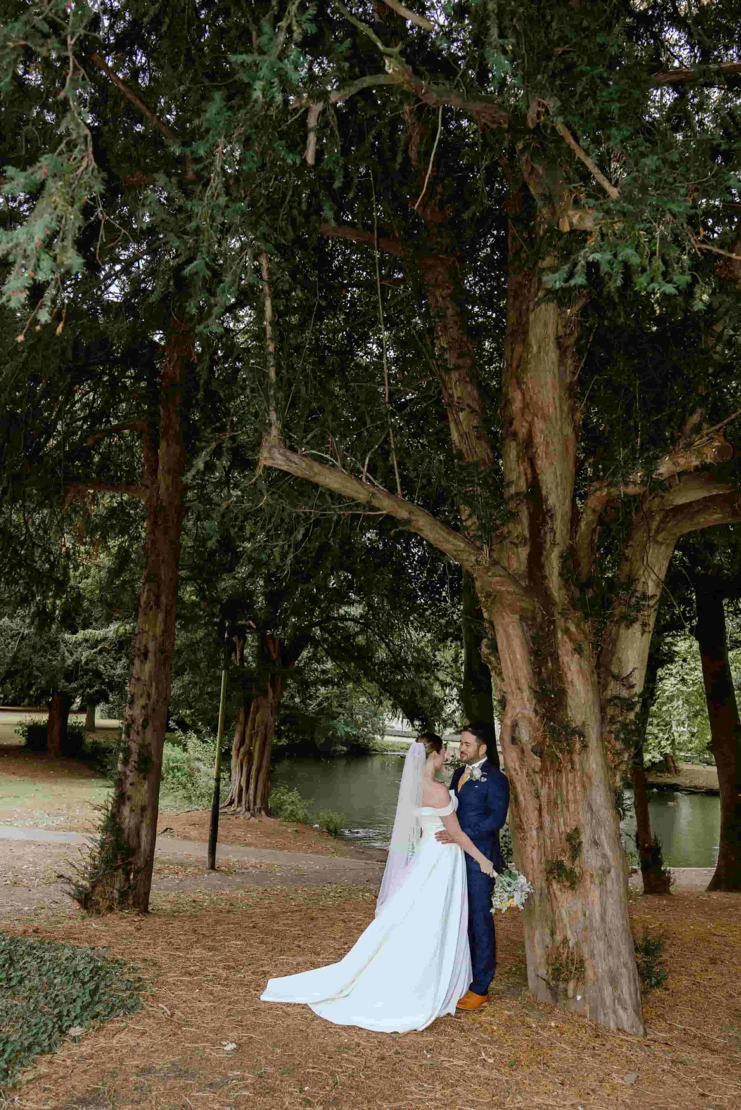 A bride and groom standing under large trees near a river, dressed in wedding attire, holding hands and gazing at each other.