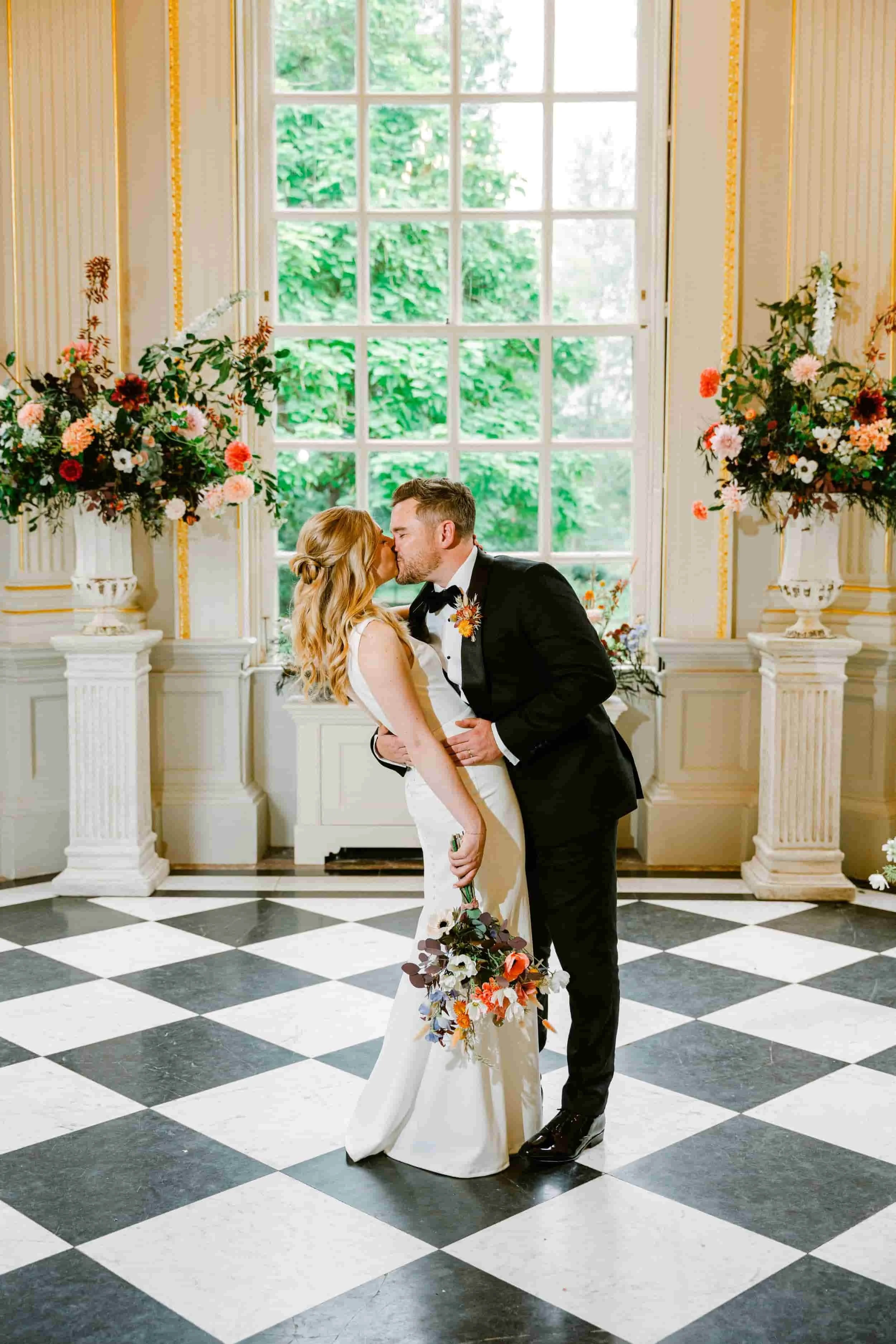 A bride and groom kiss in a decorated indoor wedding venue with black-and-white checkered floor, large window, and floral arrangements.