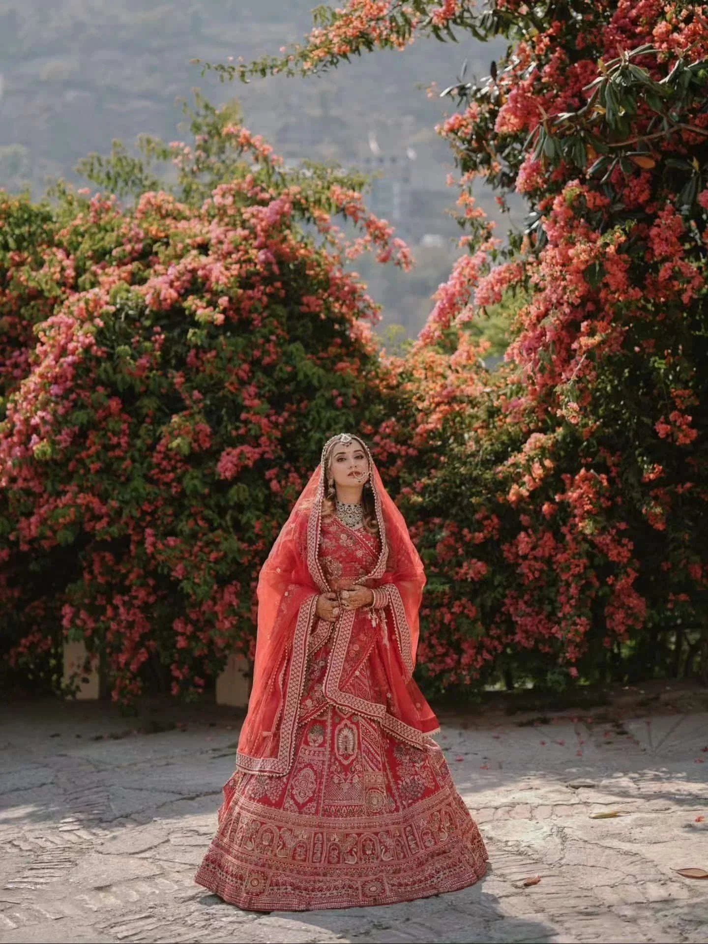 A woman dressed in traditional red embroidered attire and jewelry stands in front of lush pink flowering bushes, outdoors during daytime.