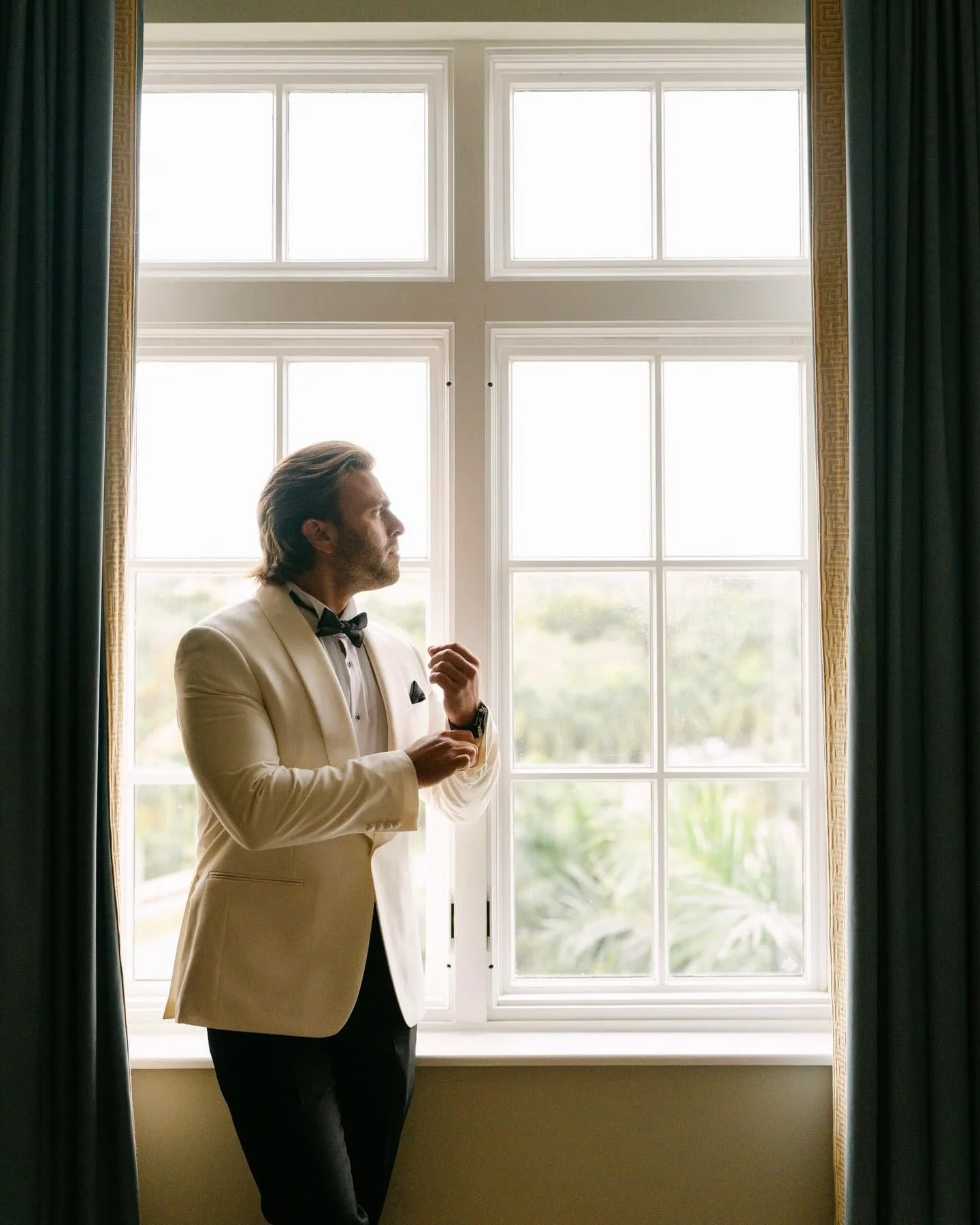A man in a white tuxedo jacket and black bow tie standing in front of a large window with green curtains, looking to the side.