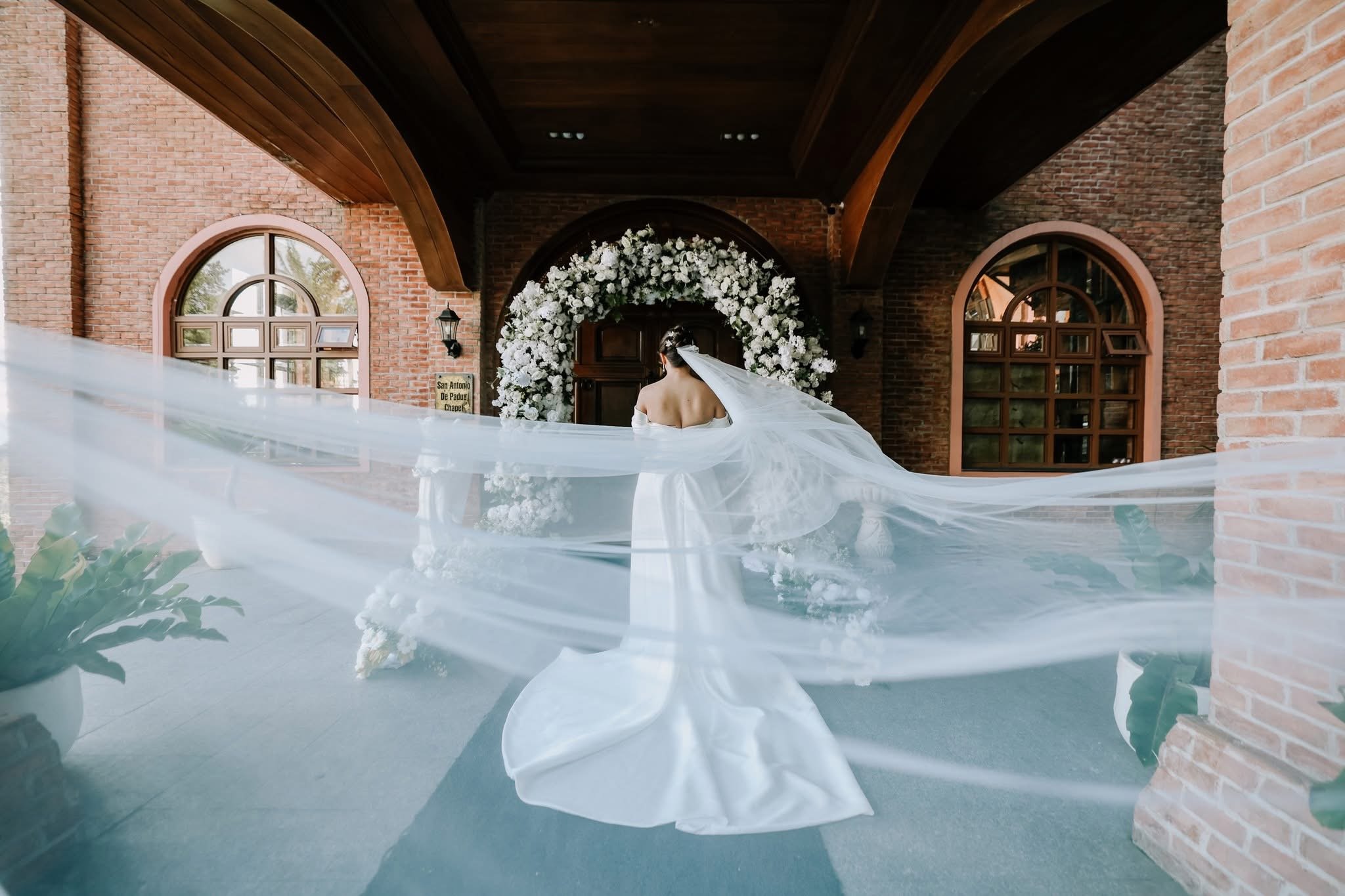 A bride in a white wedding dress and veil standing in a pool of water under a brick archway, decorated with a large white flower arrangement.
