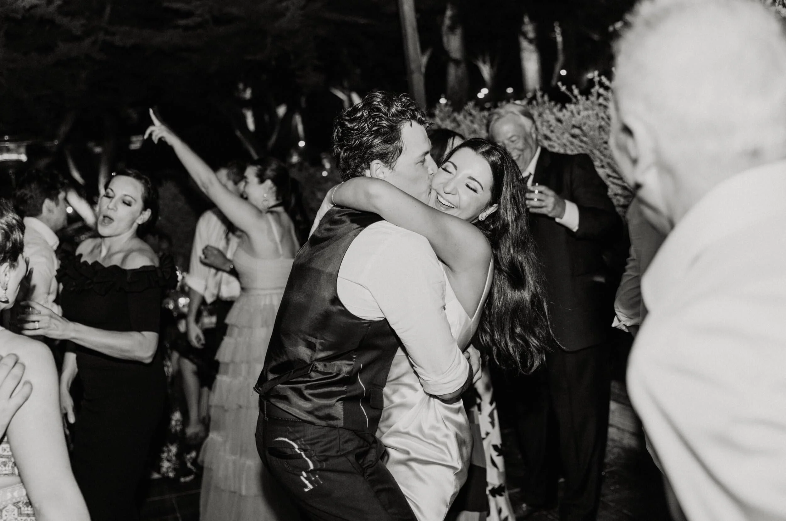 People dancing and partying at a nighttime celebration, with a man and woman embracing and kissing in the center, surrounded by other guests enjoying the event.