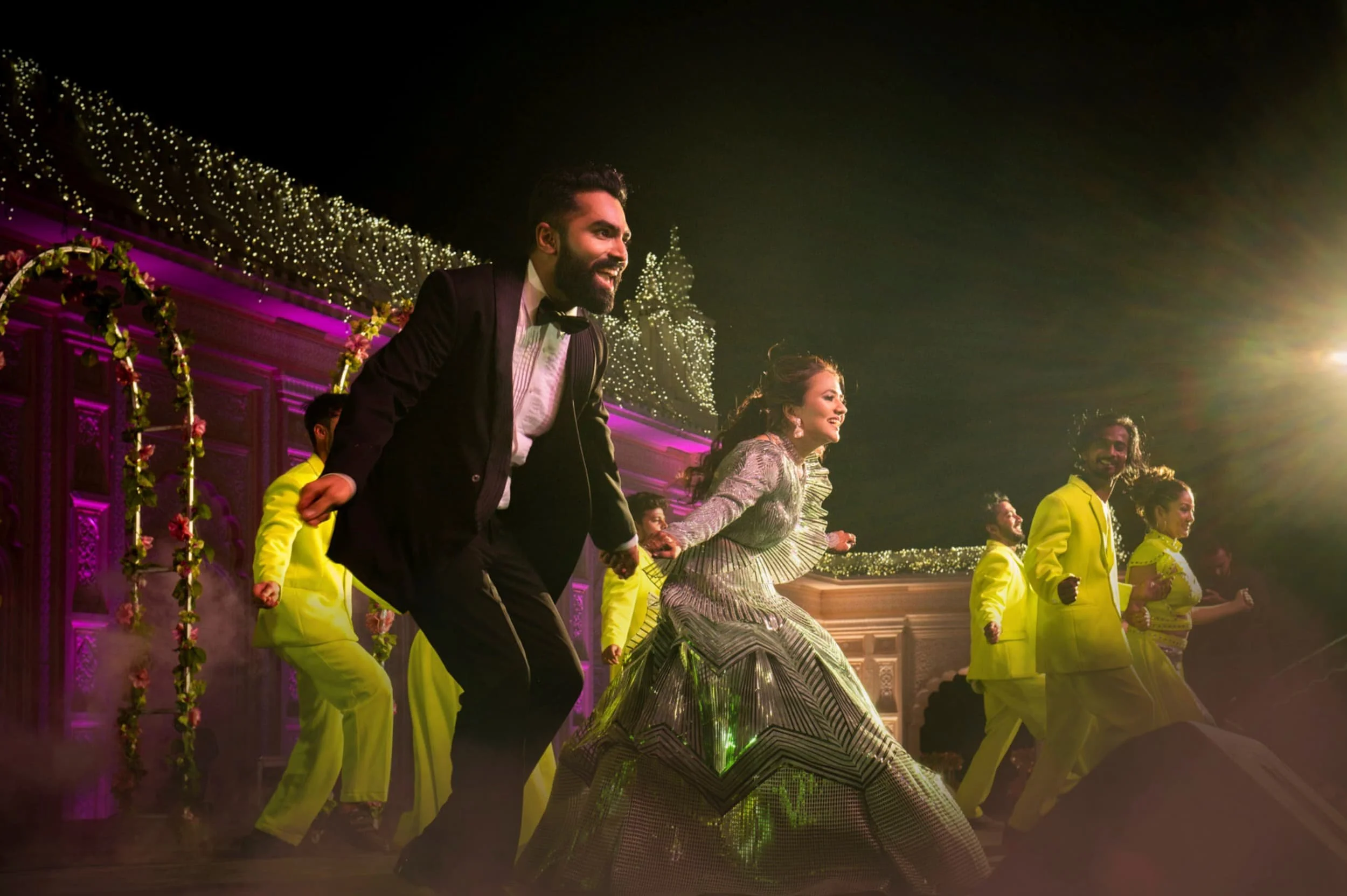 People performing a traditional dance on stage at night, decorated with string lights and floral arrangements.