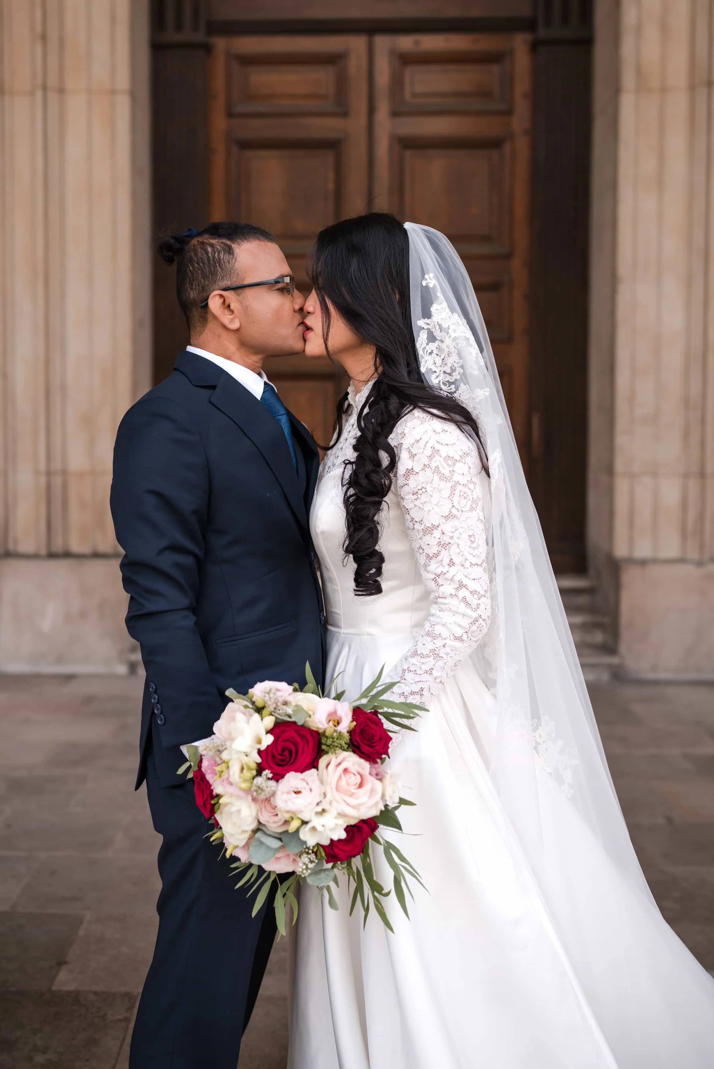 A bride and groom kiss inside a church, with the bride wearing a white lace wedding dress and a veil, and the groom in a navy suit. The bride holds a large bouquet of red, pink, and white roses and greenery.