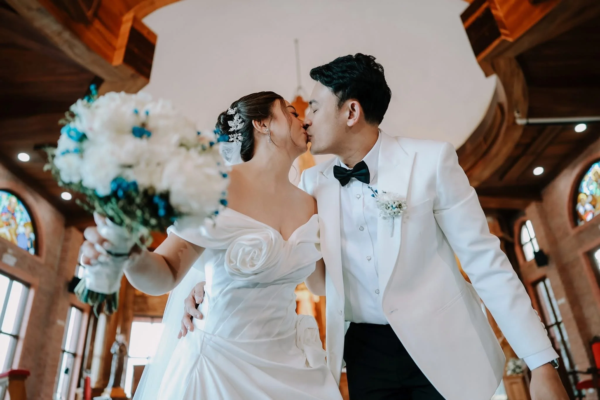 A bride and groom share a kiss inside a church with wood-paneled ceiling and stained glass windows.