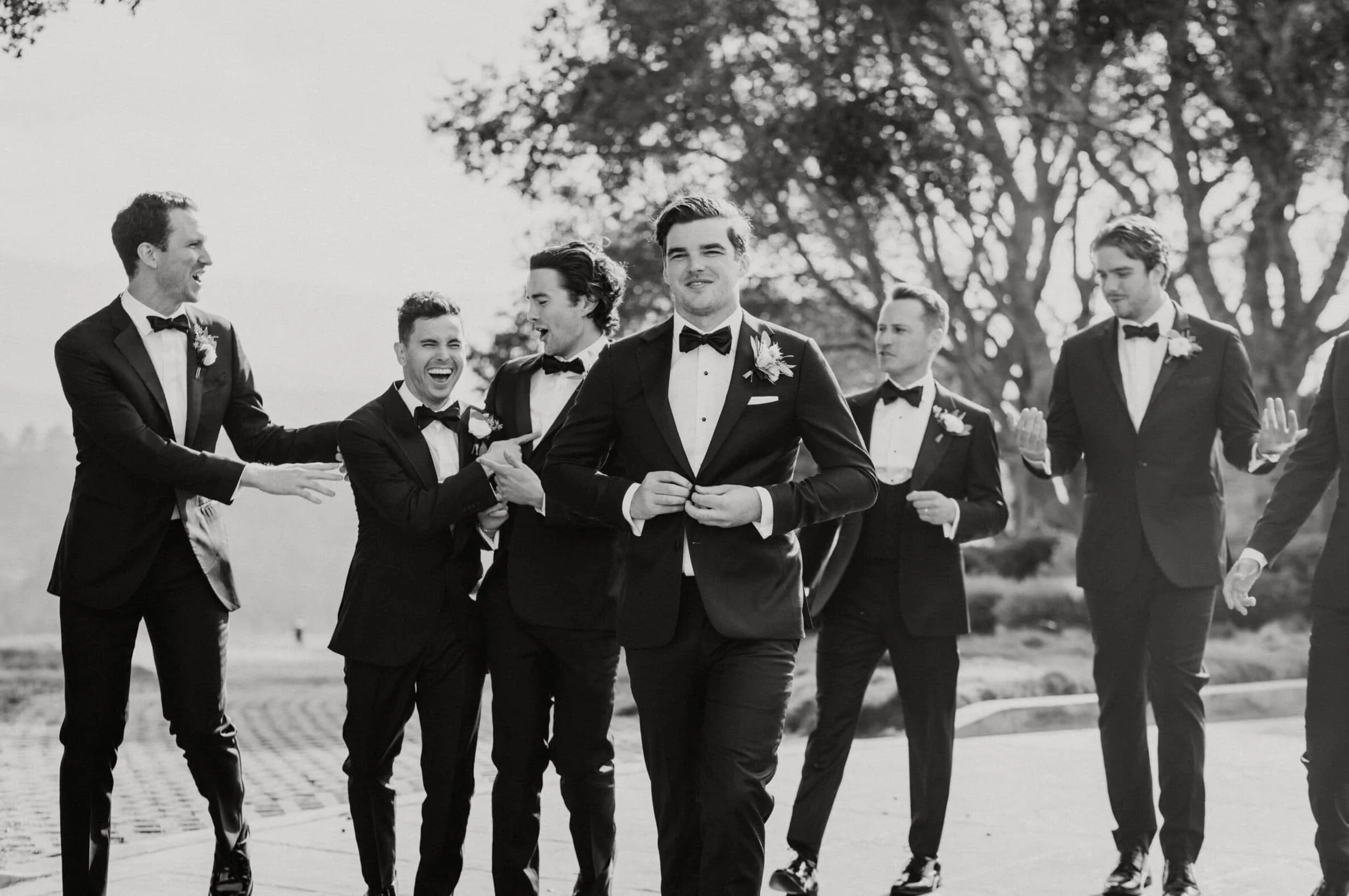 A group of men in tuxedos, some laughing and smiling, walking outdoors on a sunny day with trees in the background.