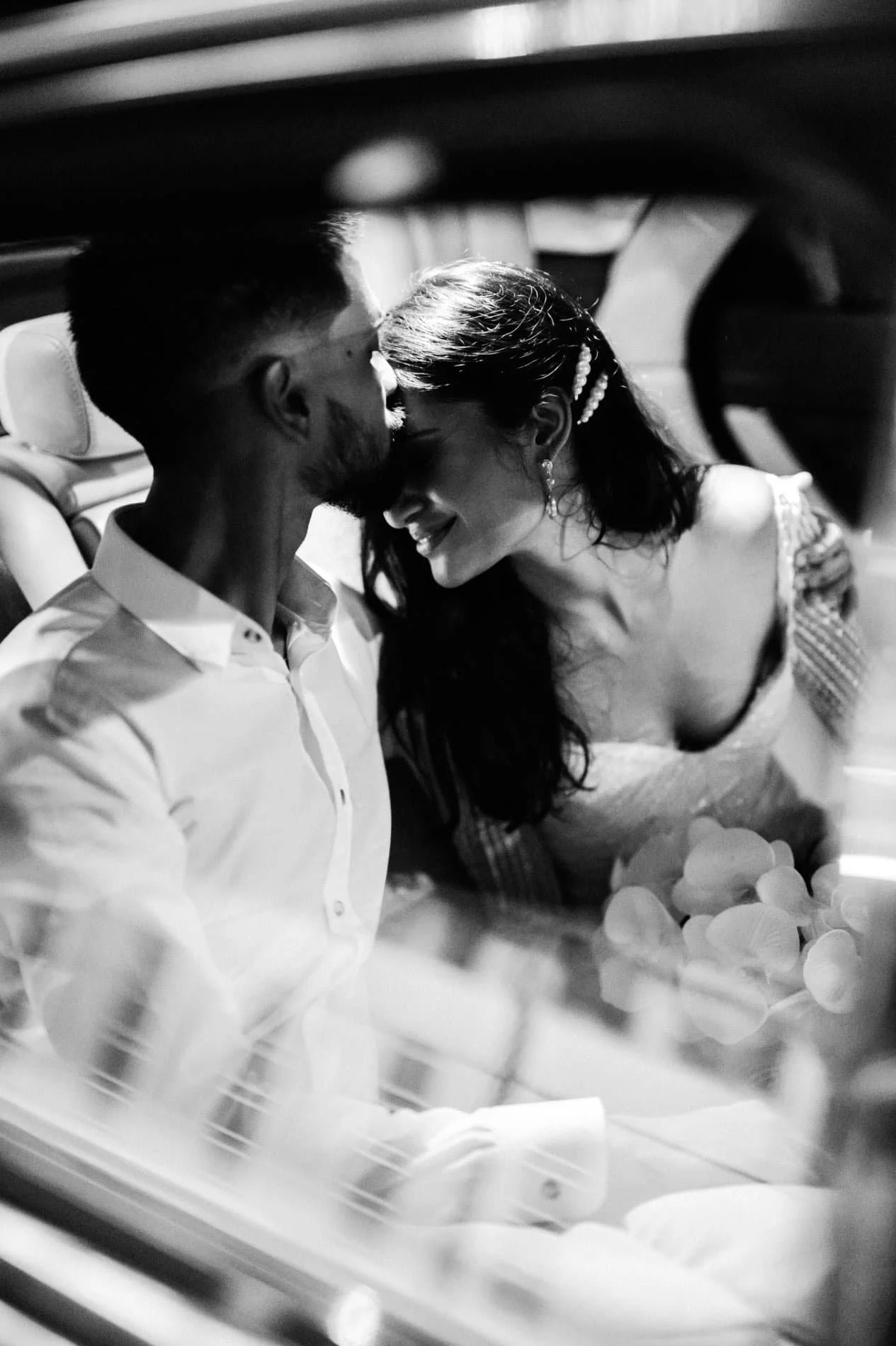 A black and white photo of a couple sharing a kiss, sitting inside a vehicle decorated with flowers, likely during a romantic or wedding moment.