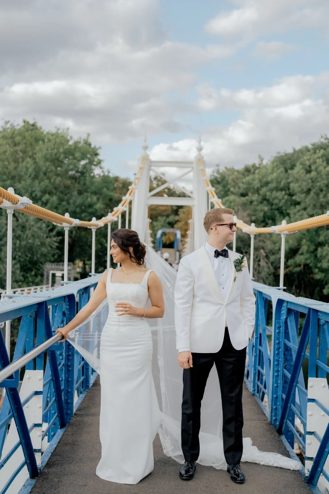 A bride in a white wedding dress and a groom in a white tuxedo with a black bow tie standing on a blue bridge with greenery in the background.