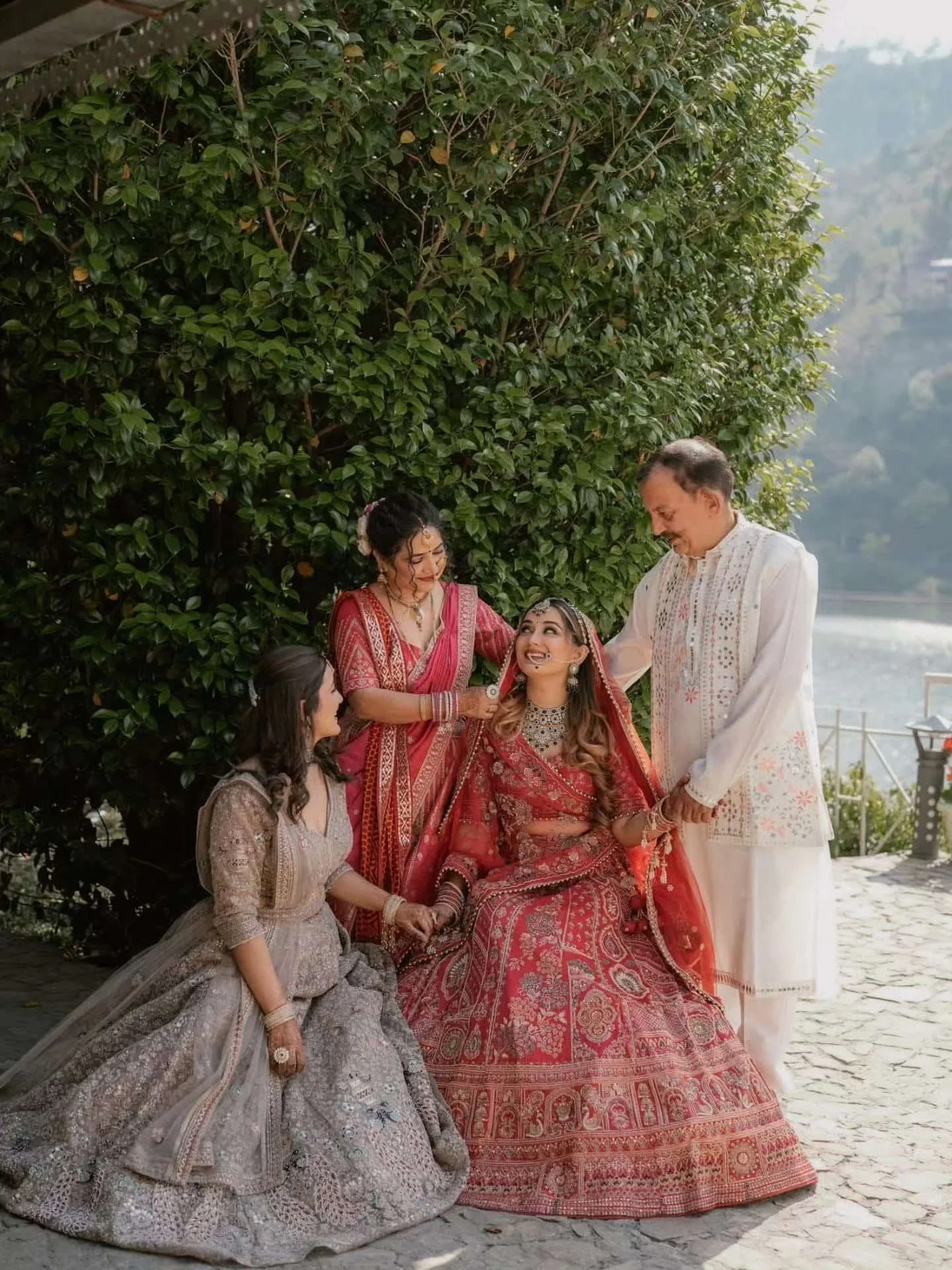 A multigenerational family of Indian ethnicity celebrating a traditional wedding outdoors near a body of water, with lush greenery in the background. The bride is dressed in a red and gold bridal outfit with extensive jewelry, seated, and smiling at 