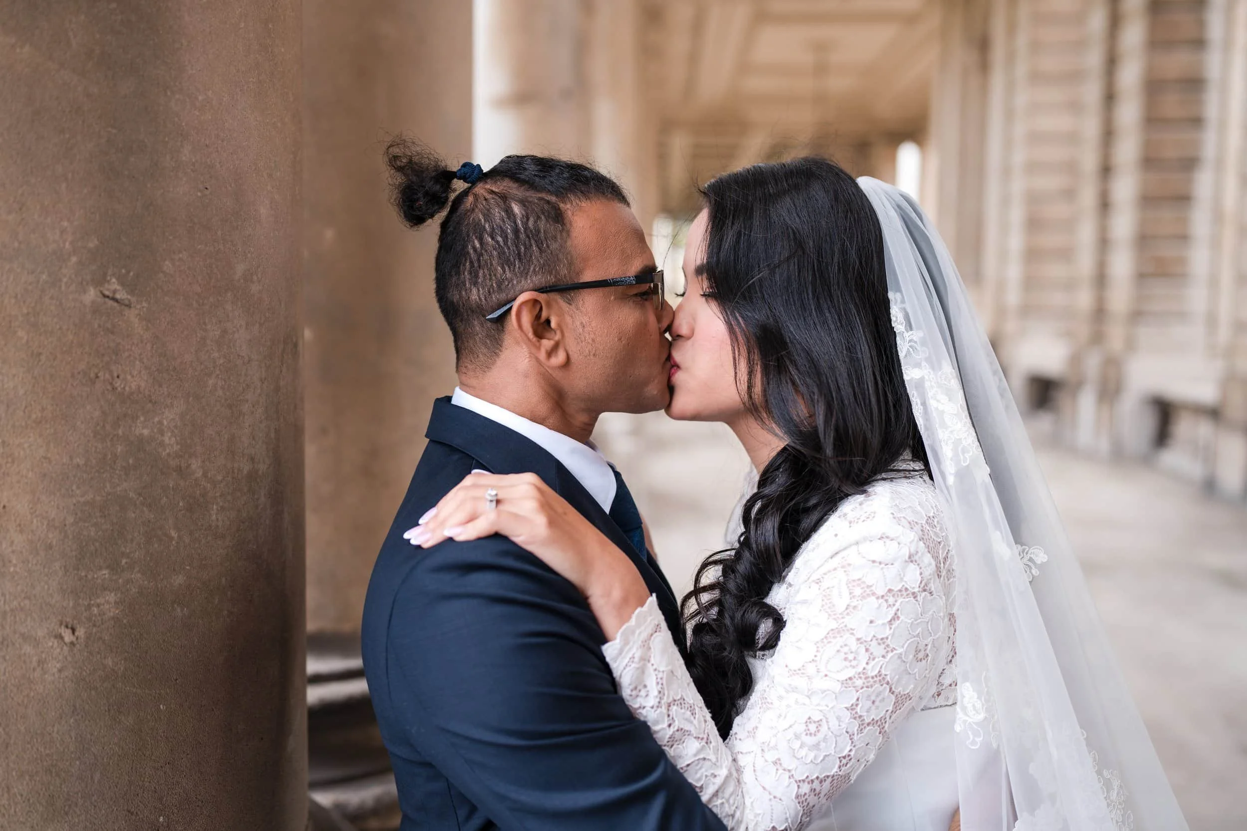 A couple shares a kiss in wedding attire, standing close against a column in an architectural corridor.