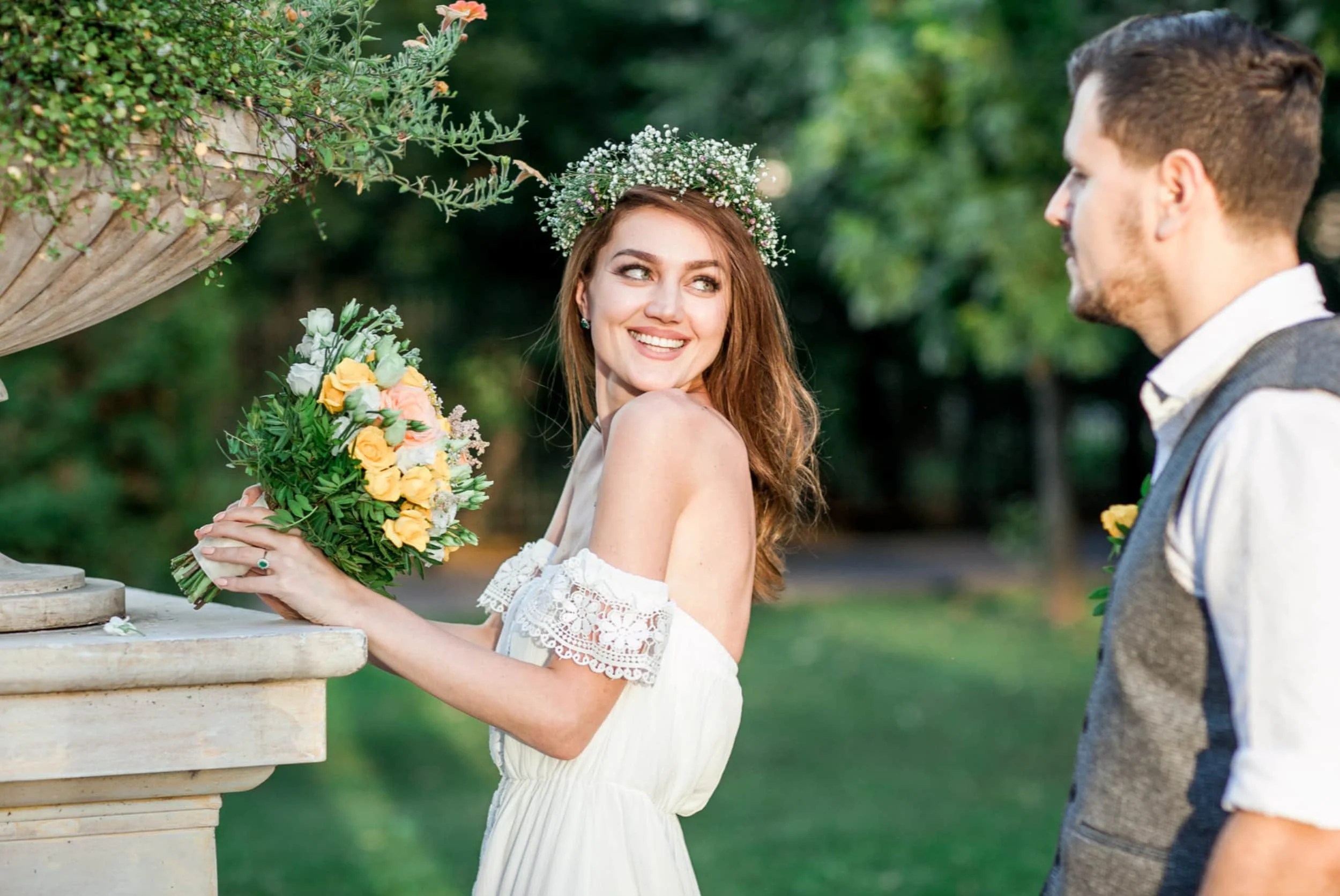 A smiling woman with long red hair wearing a white dress with lace details and a flower crown is holding a bouquet of yellow and white flowers. A man with short brown hair, wearing a vest over a white shirt, is standing nearby. They are outdoors with