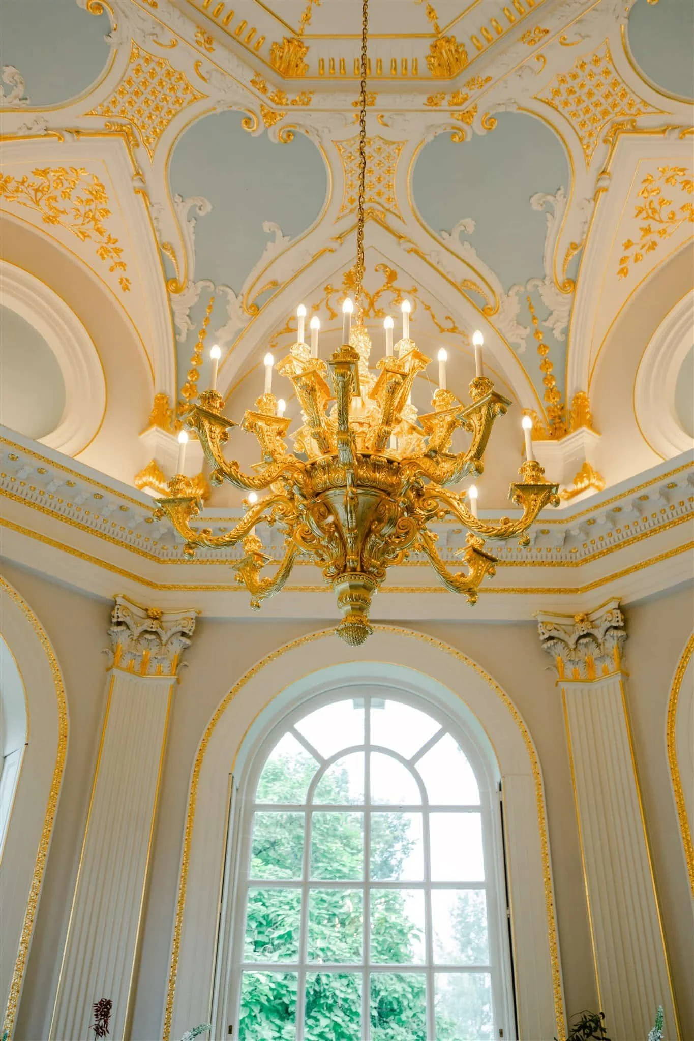 Elegant room with a gold chandelier hanging from a ceiling decorated with gold accents, molding, and ornate designs. Large arched window allows natural light to enter, with green trees visible outside.