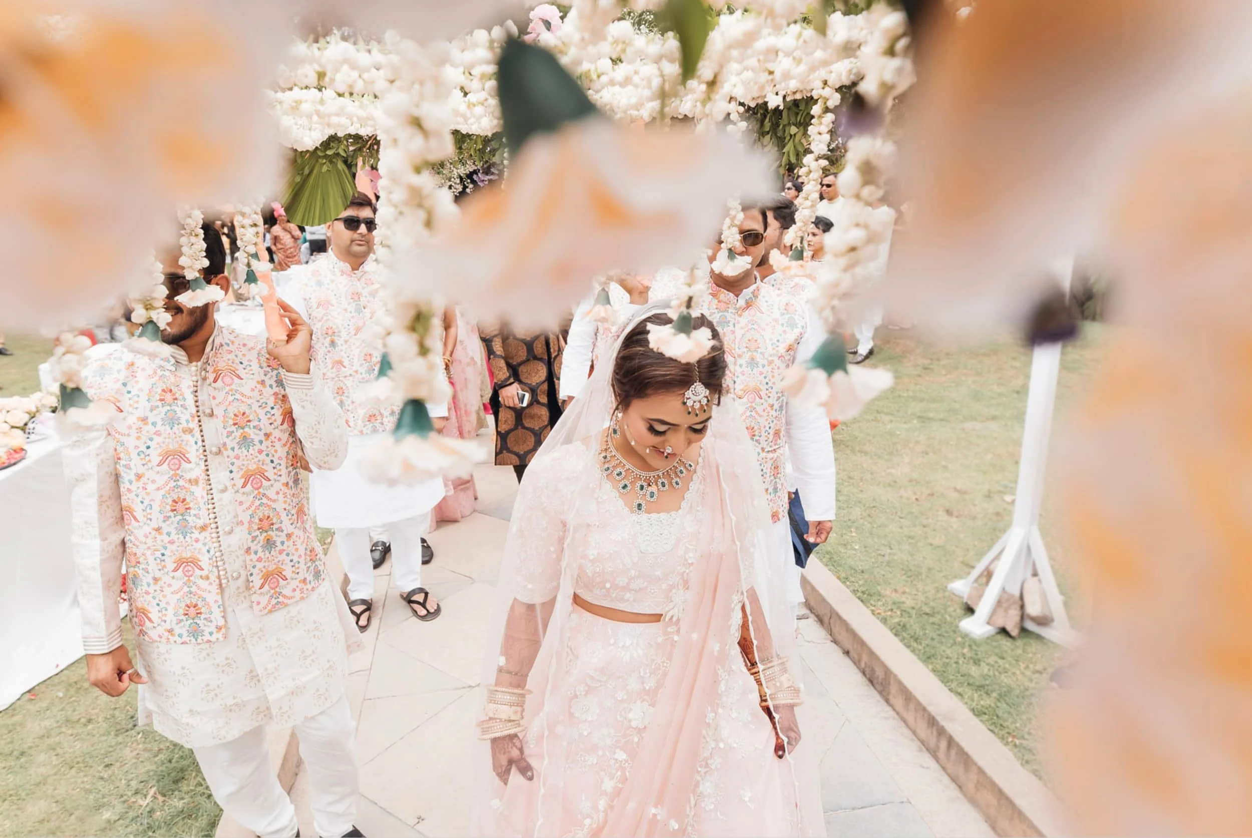 A wedding procession outdoors, seen through a decorative floral arch, featuring a bride in traditional attire and jewelry, surrounded by men in patterned fits, some wearing sunglasses, under a cloudy sky.