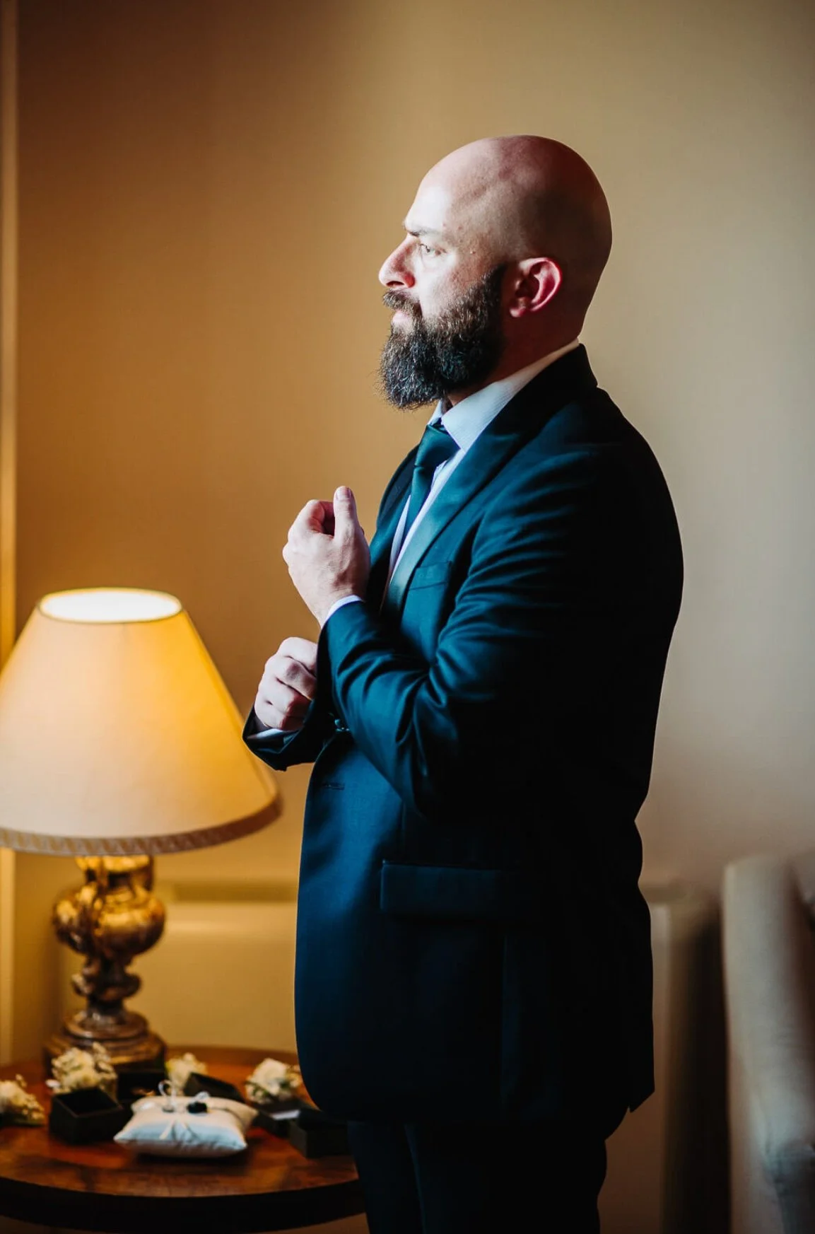 A man with a beard and bald head in a suit adjusting his cufflink in a warmly lit room, with a table holding jewelry and a lamp in the background.