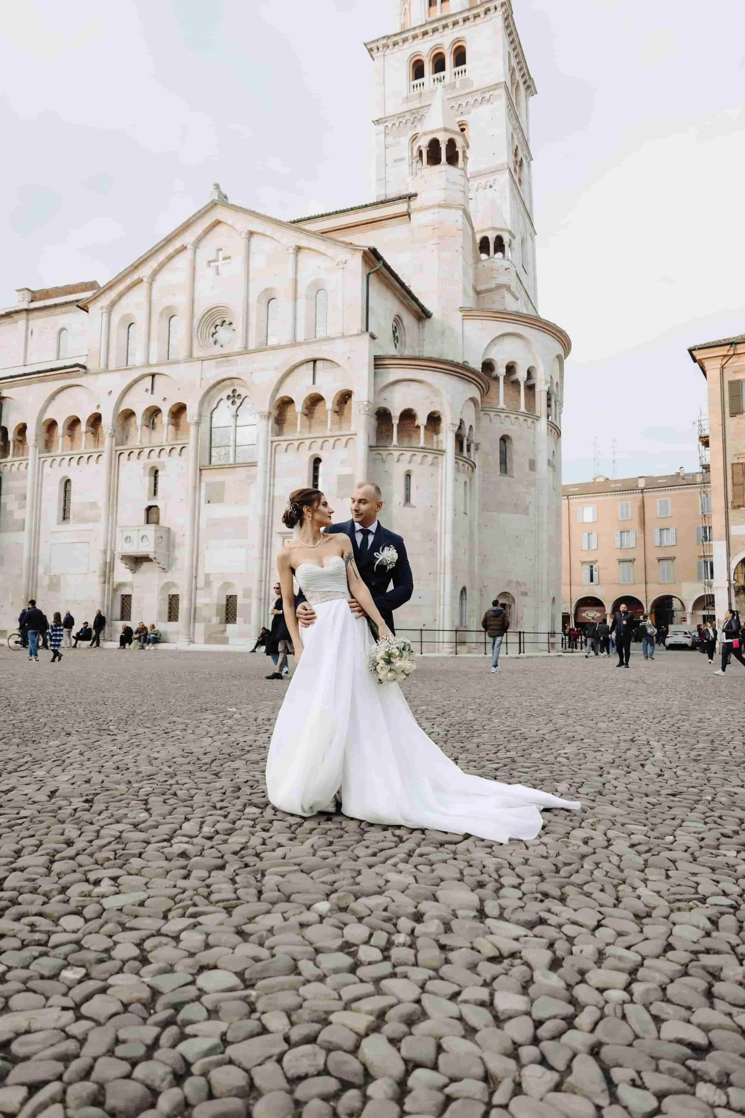 A bride and groom in wedding attire standing on a cobblestone square in front of a historic church with a tall bell tower, surrounded by people.