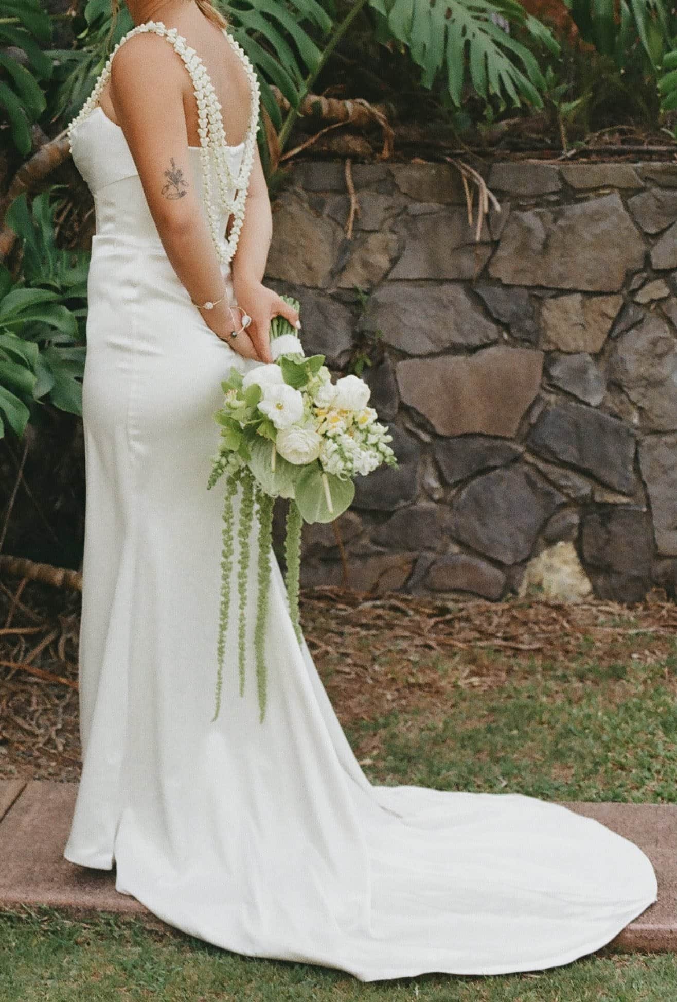 A woman in a white wedding dress holding a bouquet of white flowers standing outdoors in front of a stone wall and green plants.