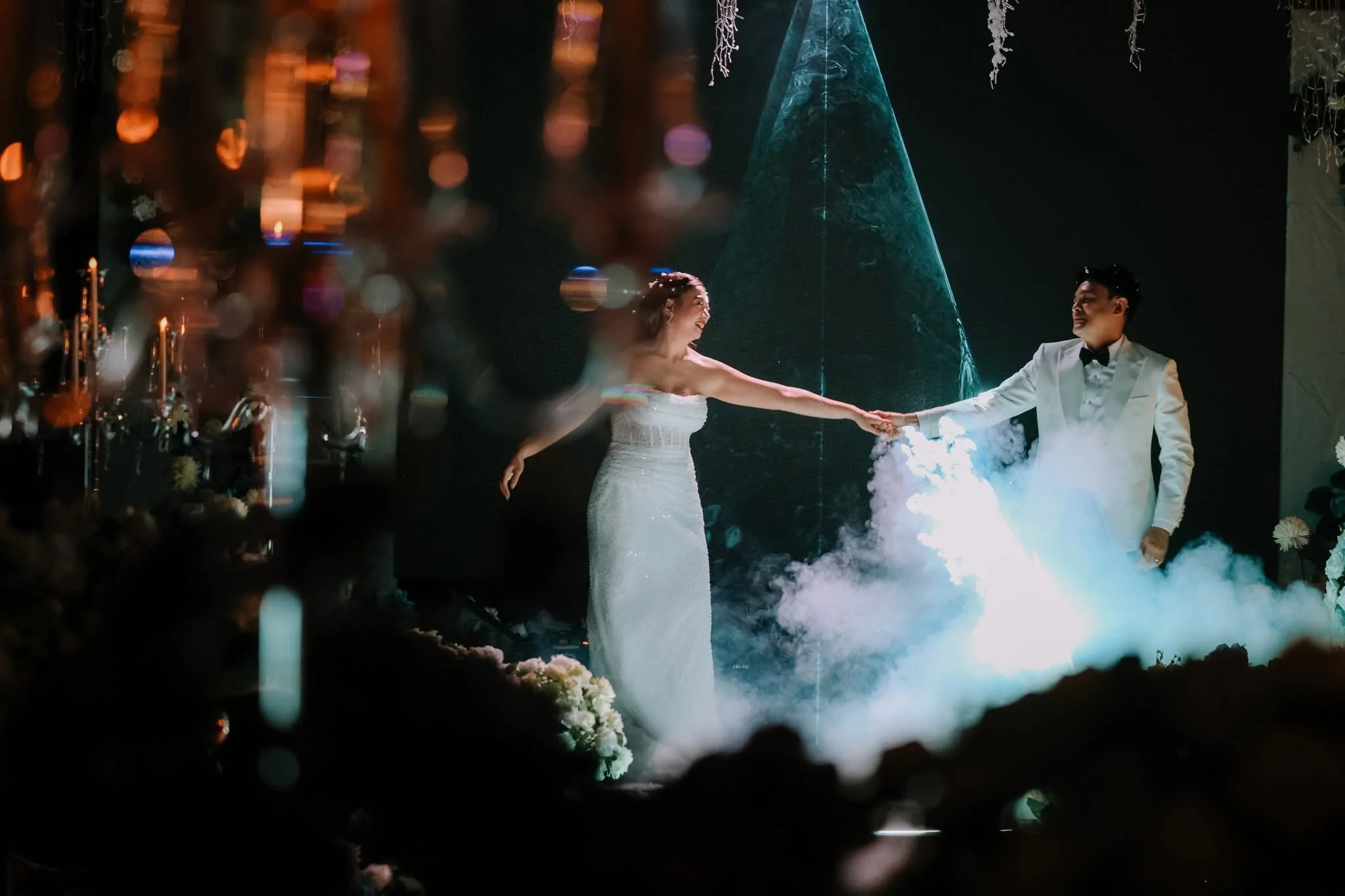 A bride and groom holding hands during their wedding reception, surrounded by fog and vibrant lighting, with a blue water fountain in the background.