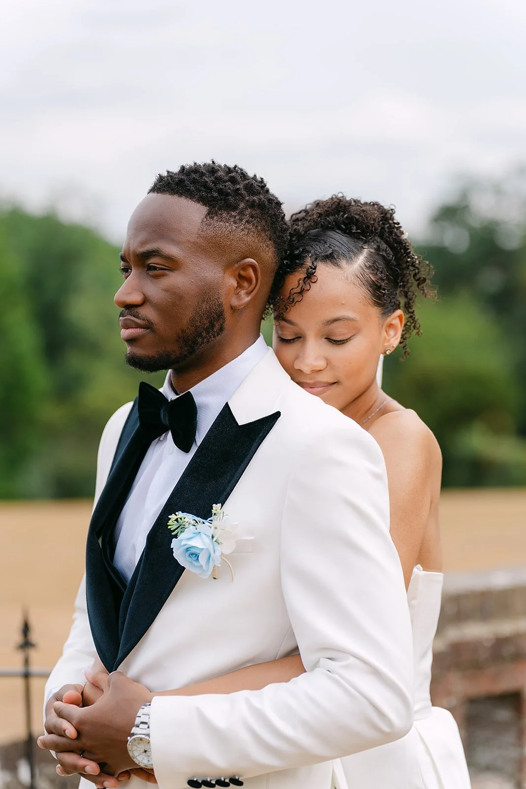 A bride and groom outdoors on their wedding day, with the bride standing behind the groom, her arms around his waist, and her eyes closed.