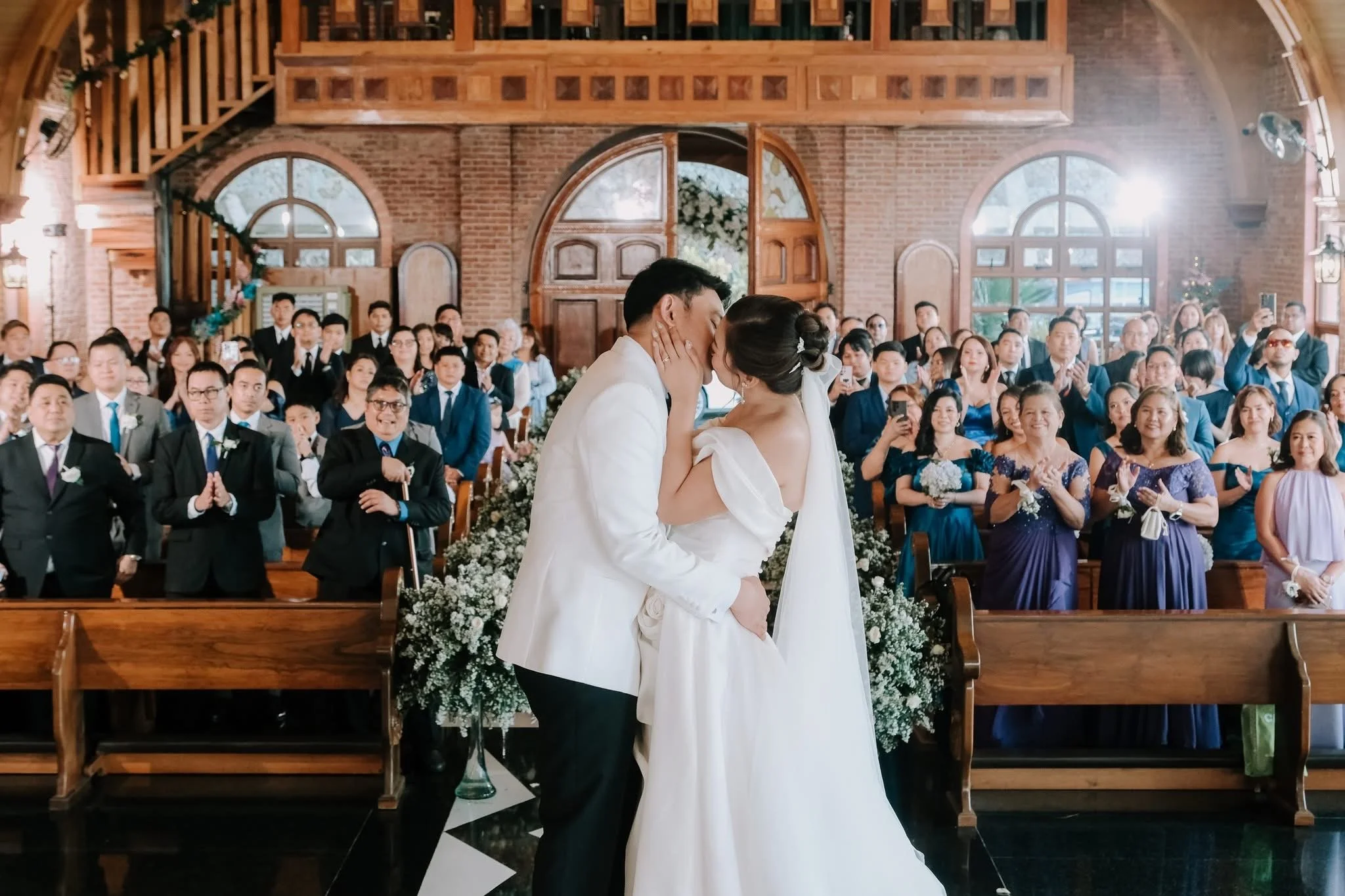 A bride and groom sharing a kiss during their wedding ceremony inside a church filled with guests in formal attire, with wooden architectural features and large windows in the background.