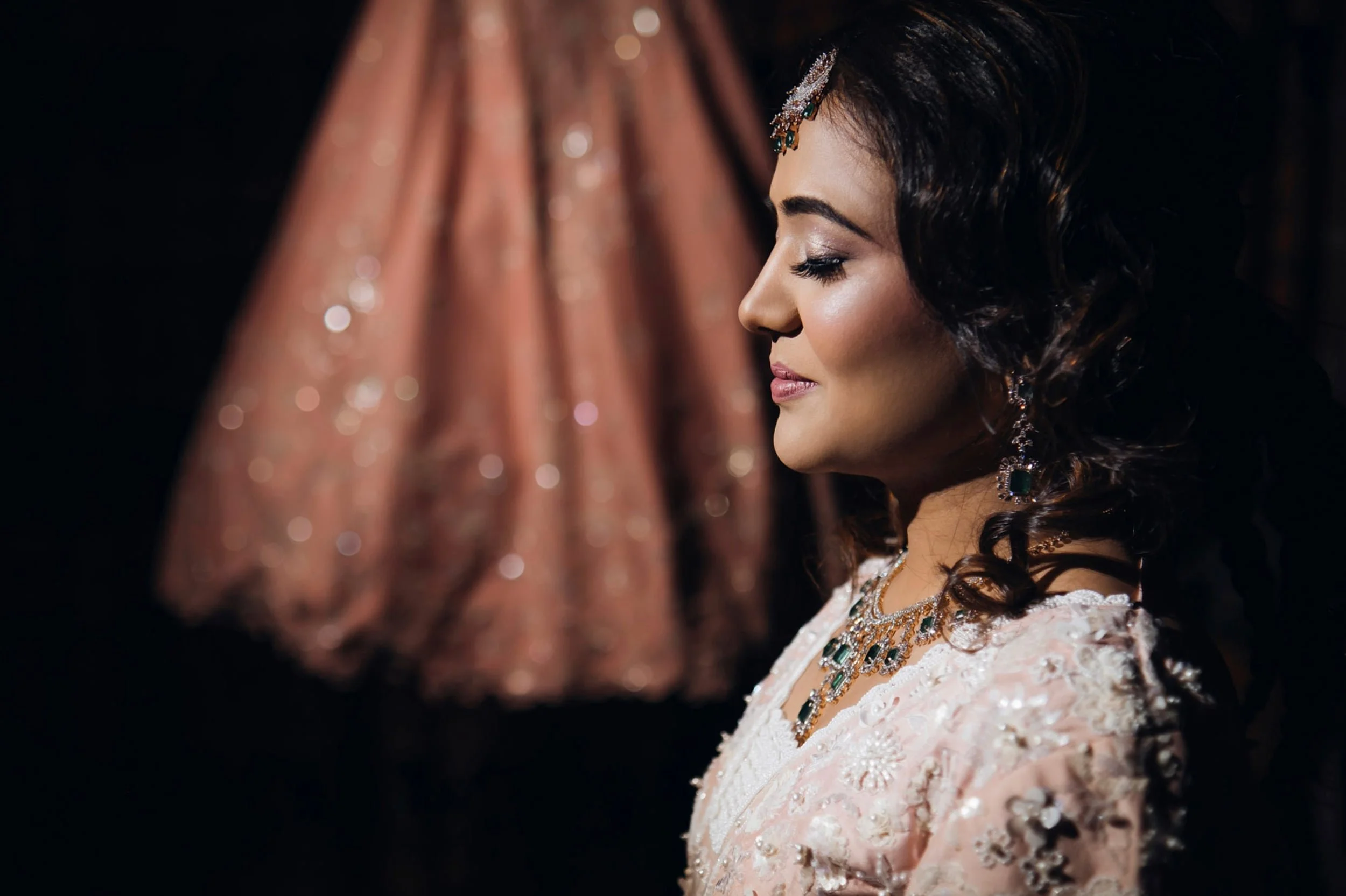 A woman dressed in traditional South Asian bridal attire, with elaborate jewelry and styled hair, standing in profile against a dark background. A blurred, ornate pink outfit is visible in the background.