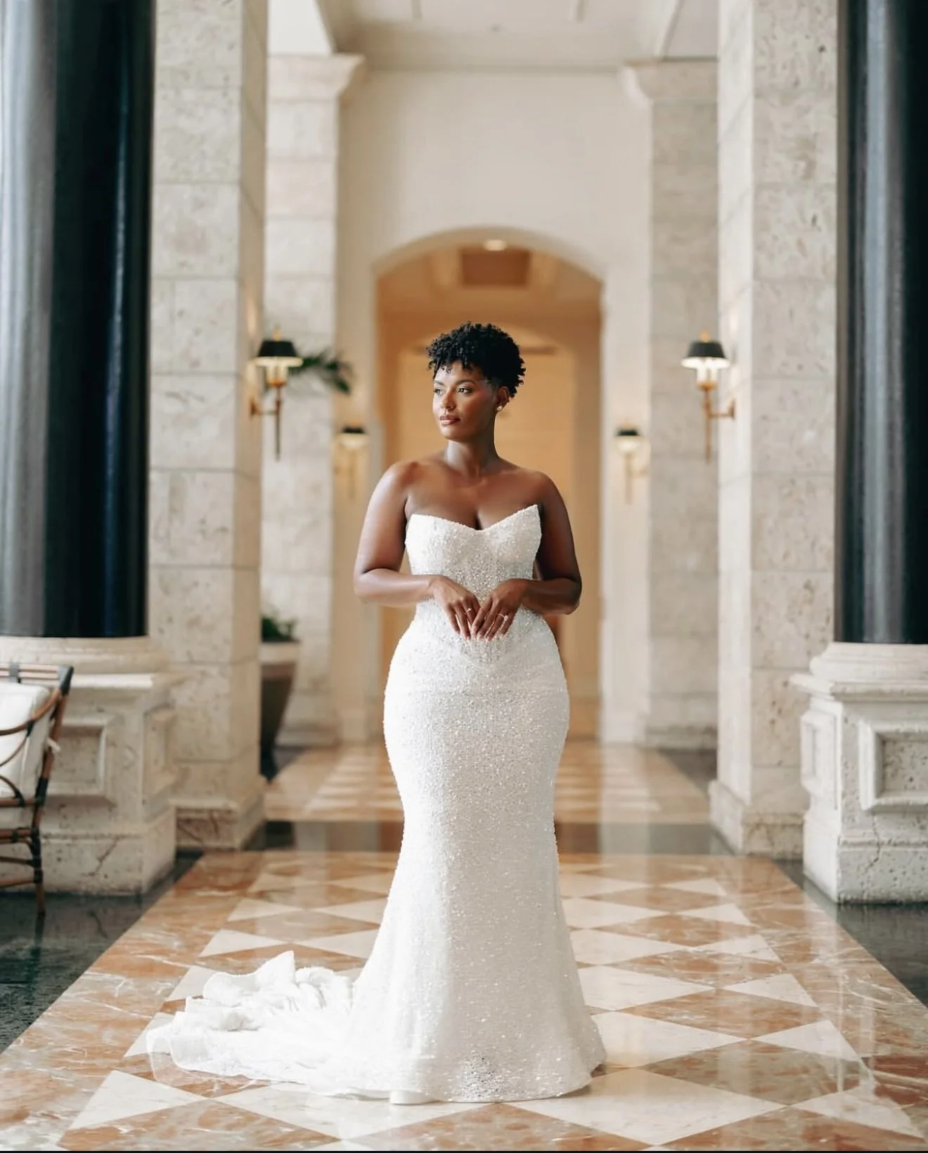 A woman in a strapless white sequin wedding gown standing in an elegant, marble-floored hall with stone walls and wall sconces.