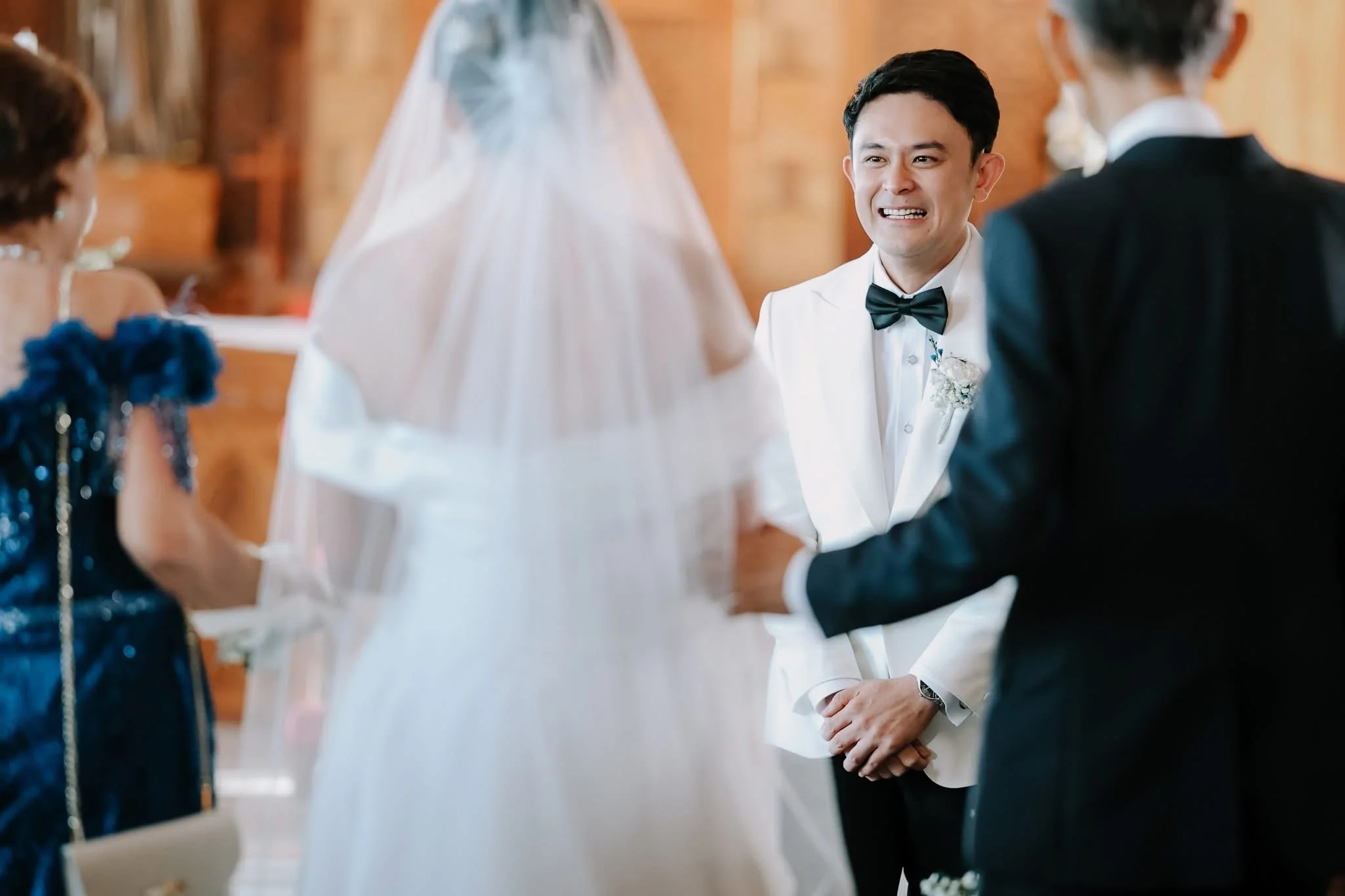 A groom in a white tuxedo and a bride in a white wedding dress holding hands during a wedding ceremony, with a priest and a woman in a blue dress in the background.
