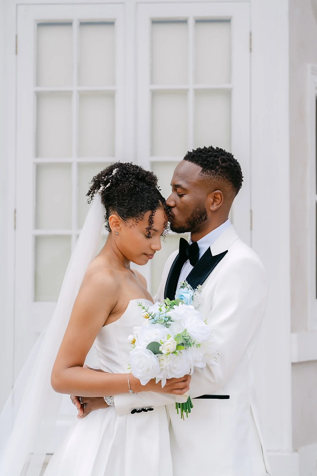 A bride and groom sharing a tender moment at their wedding, the groom kissing the bride on the forehead while she holds a bouquet of white flowers.