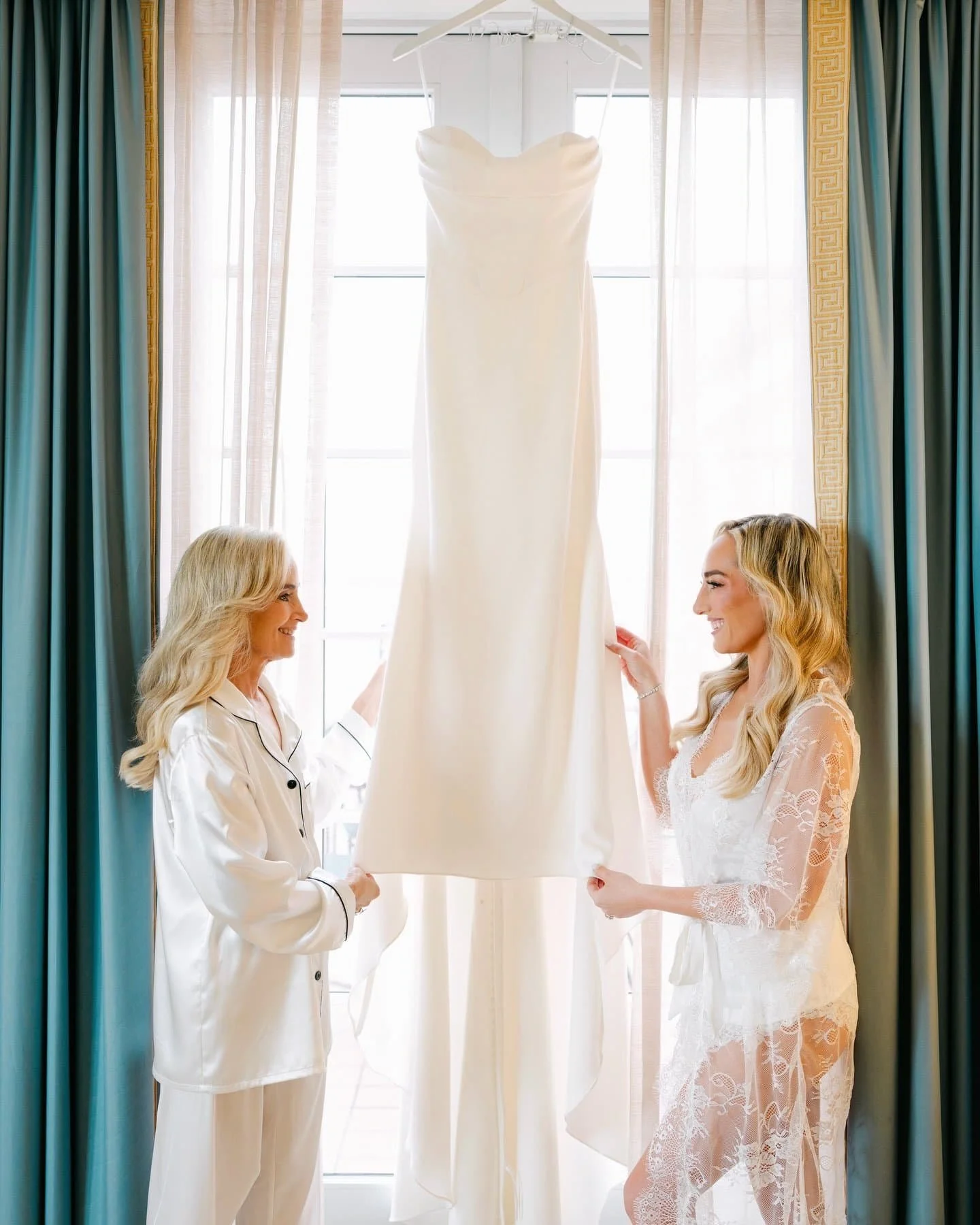 Two women, one in pajamas and one in a lace bridal robe, holding a wedding dress between them in a bright room with large windows and curtains.