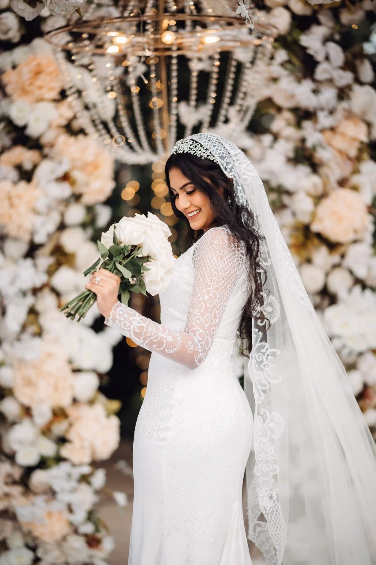 A bride in a white wedding dress holding a bouquet of white flowers, standing in front of a floral backdrop with hanging lights and chandeliers.