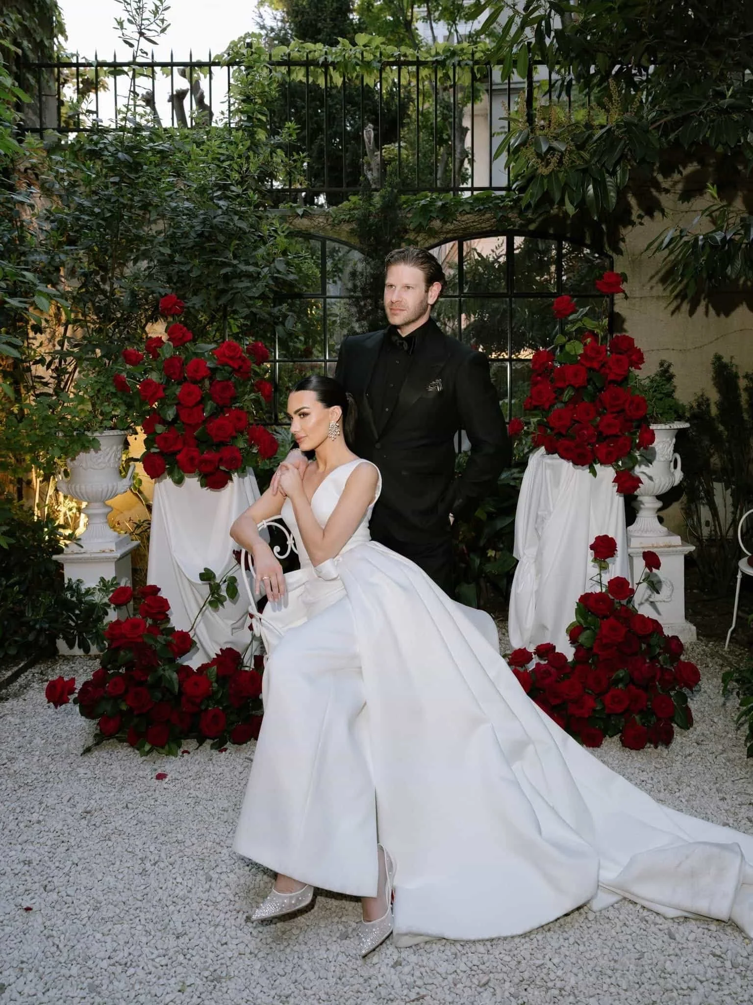A bride in a white wedding gown sitting in front of a backdrop of red roses and greenery, with a groom standing behind her in a black suit. The scene is outdoors, decorated with large white vases filled with red roses, set on a gravel ground.