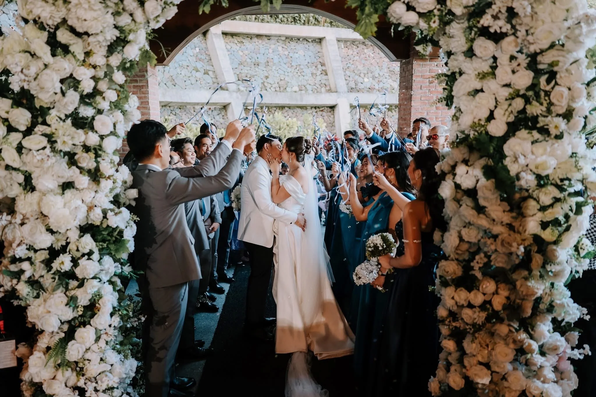 A wedding ceremony with a couple kissing under an arch decorated with white flowers, surrounded by family and friends celebrating.
