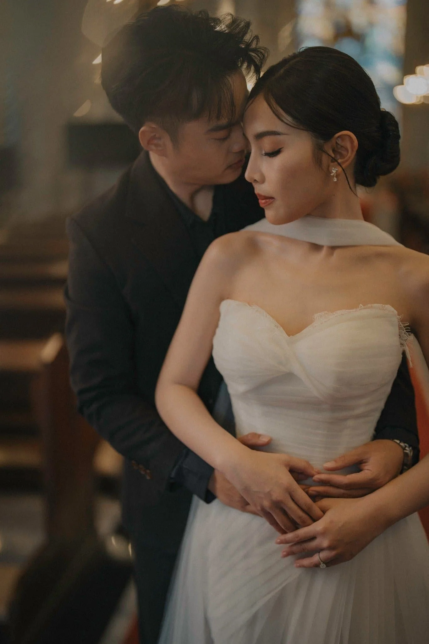 A couple dancing closely at a wedding reception, with the man gently holding the woman's waist and the woman resting her hand on his shoulder. The woman is wearing a white dress with off-shoulder sleeves, and the man is in a dark suit. They have thei