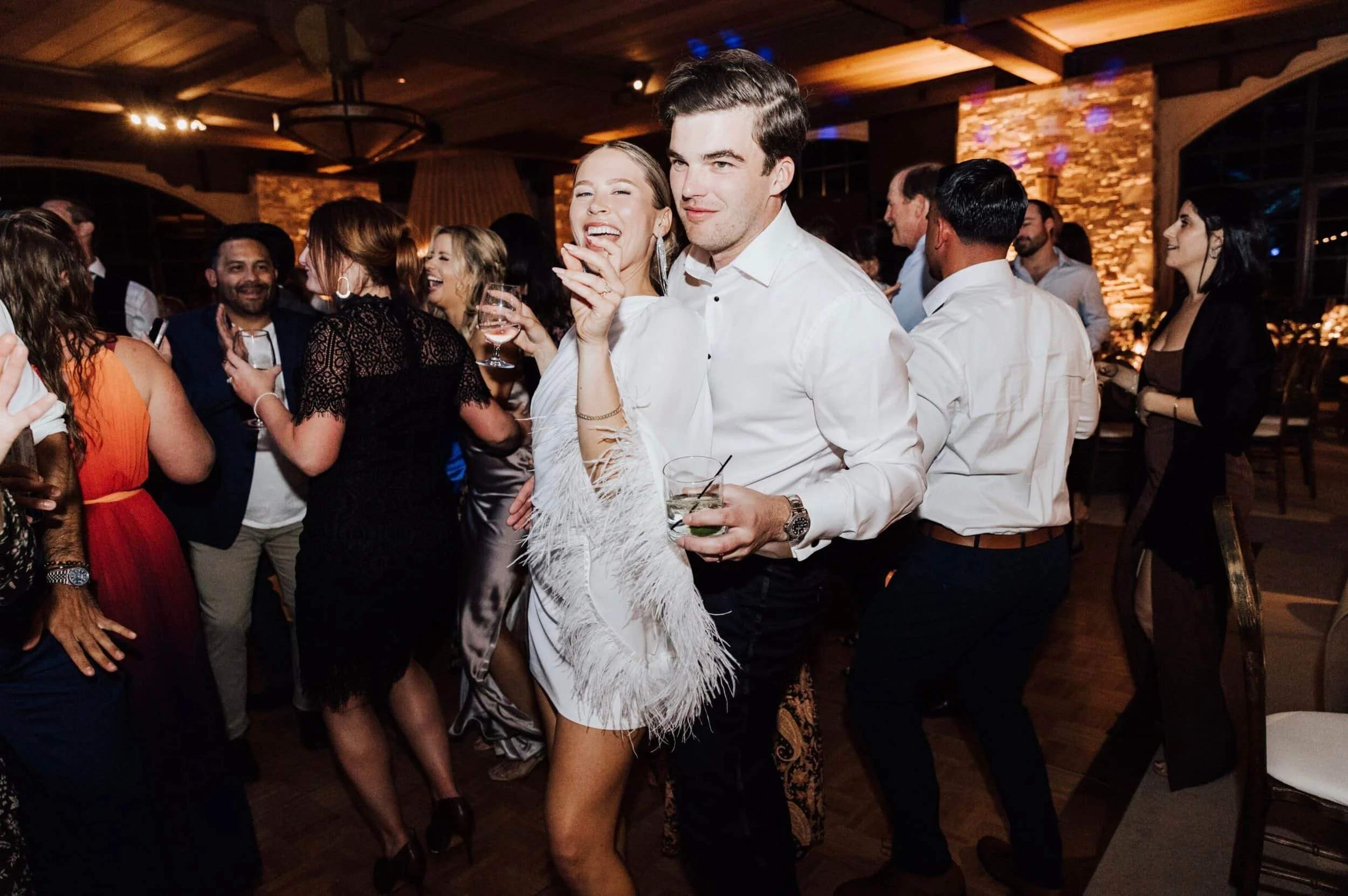People dancing and socializing at a lively indoor party or celebration, with a woman in a white dress and a man in a white shirt in the foreground.