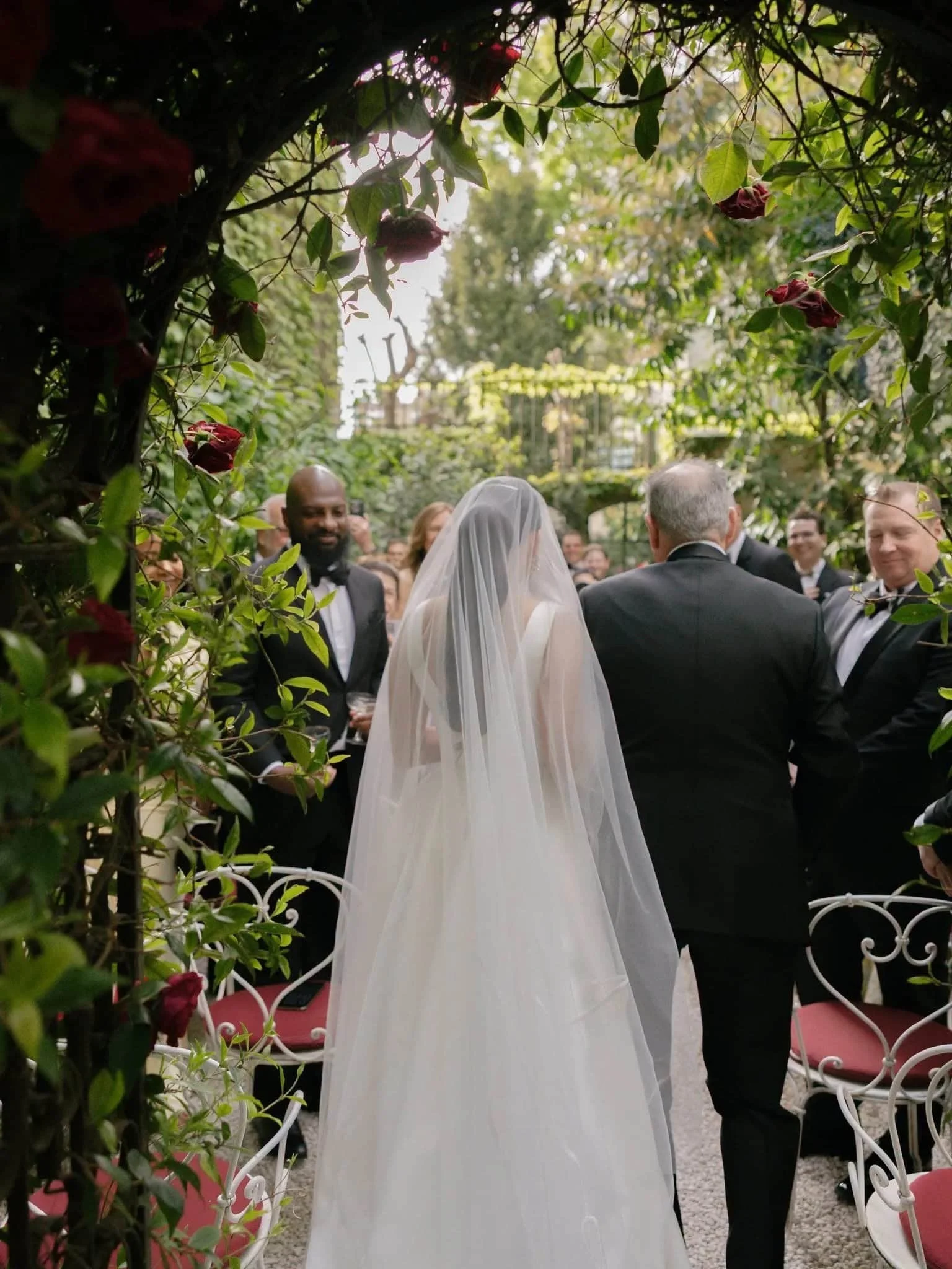 Bride and groom standing together during a wedding ceremony outdoors, surrounded by guests and lush greenery with flowers.