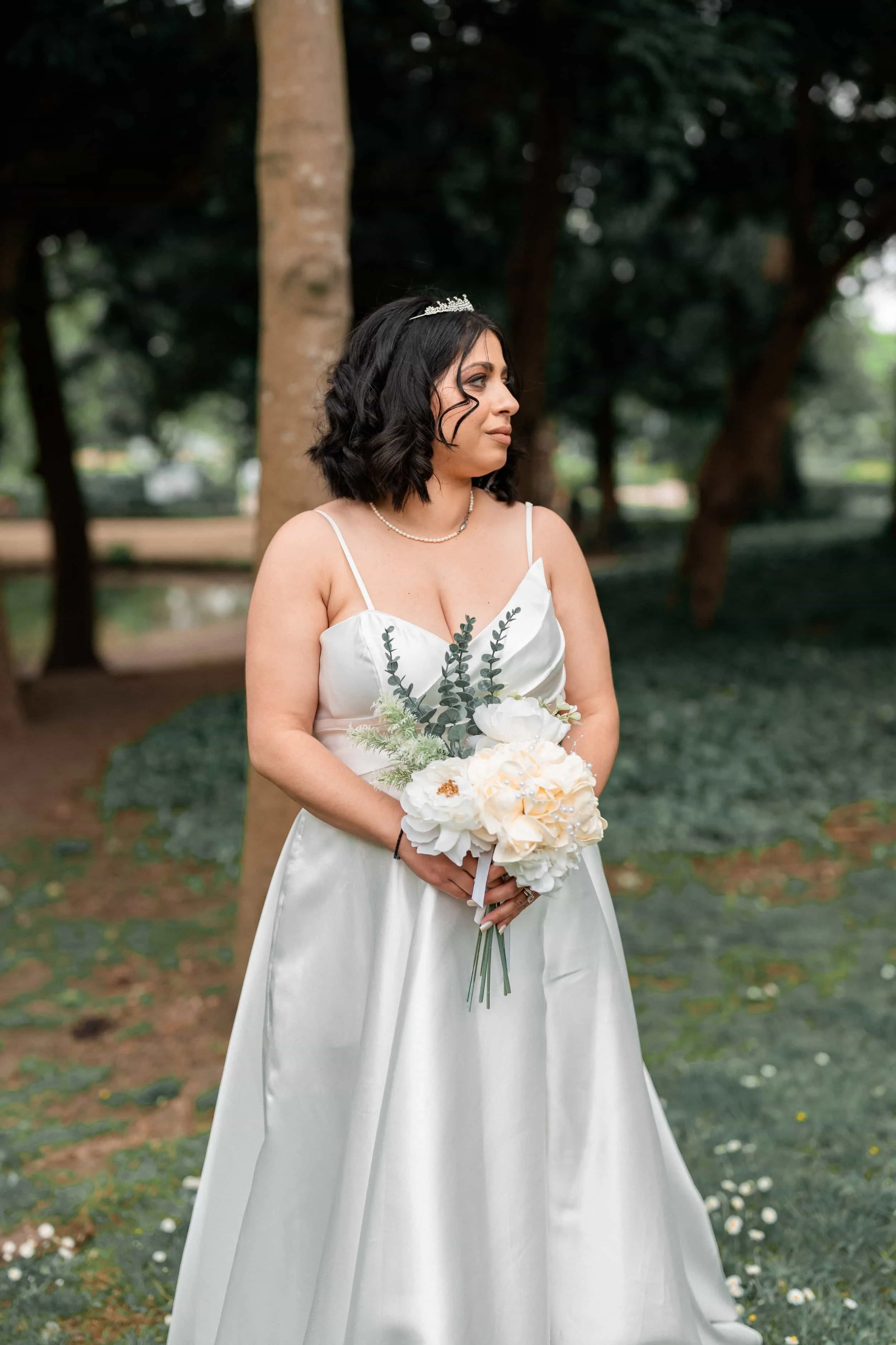 A woman in a white satin wedding dress holding a bouquet of white flowers and greenery, standing outdoors in a park with trees in the background, wearing a Tiara and pearl necklace, looking to the side.