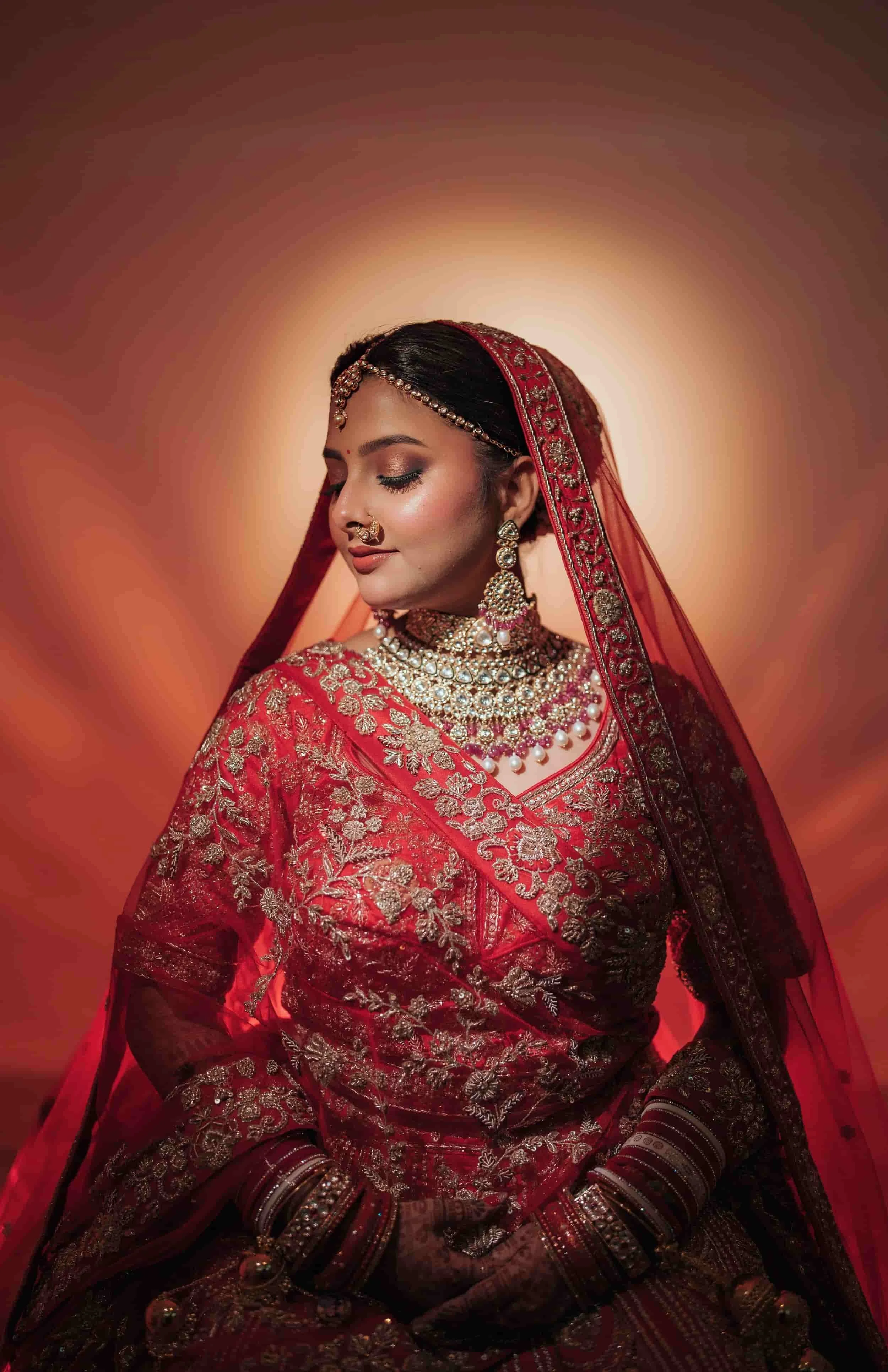 A woman dressed in traditional Indian bridal attire, with a red and gold embroidered saree, ornate jewelry, and henna-covered hands, posing with her eyes closed against a warm, gradient background.