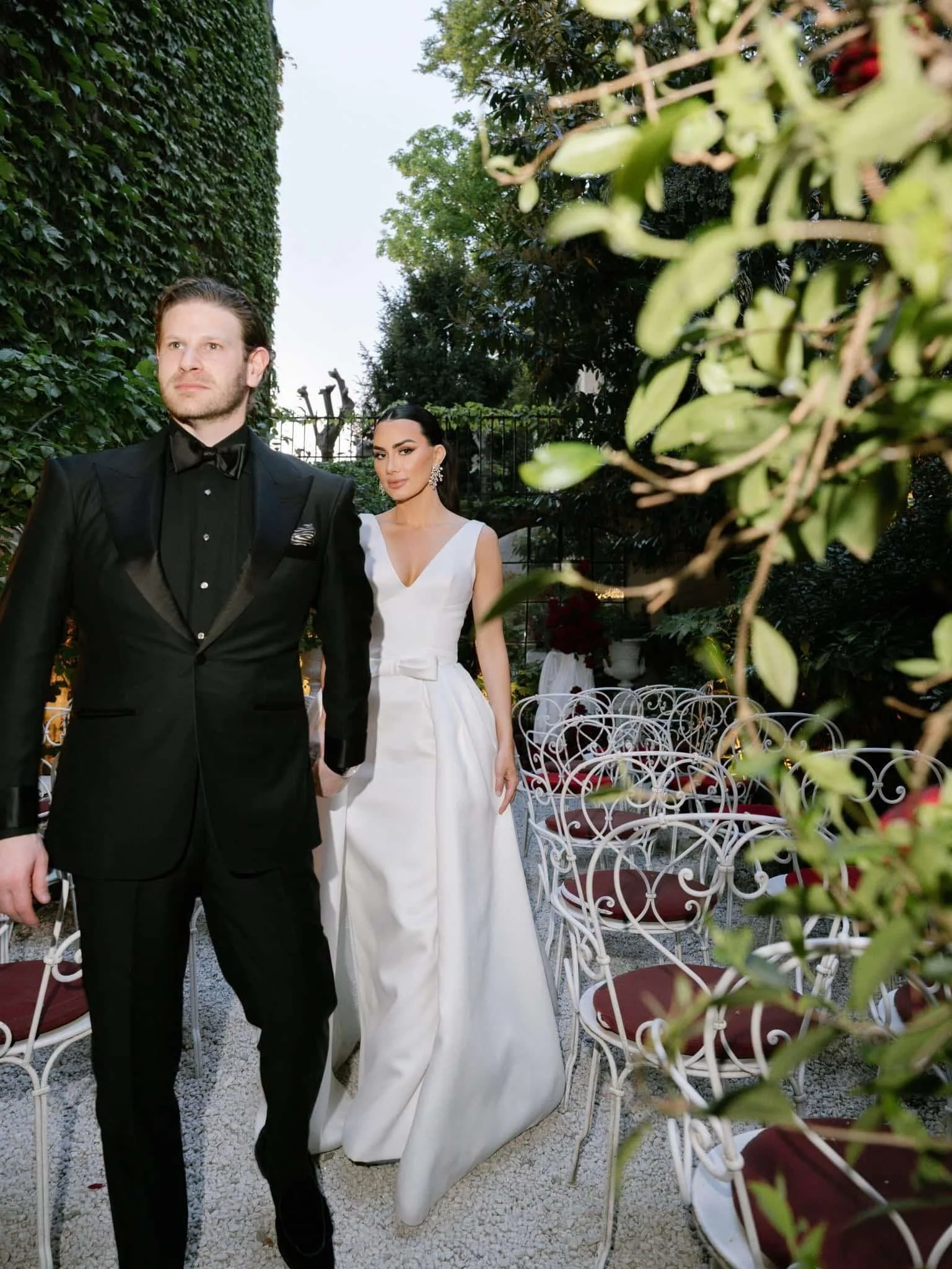 A man and woman dressed in formal attire standing outdoors among white wrought iron chairs and green foliage, with a fence and trees in the background.