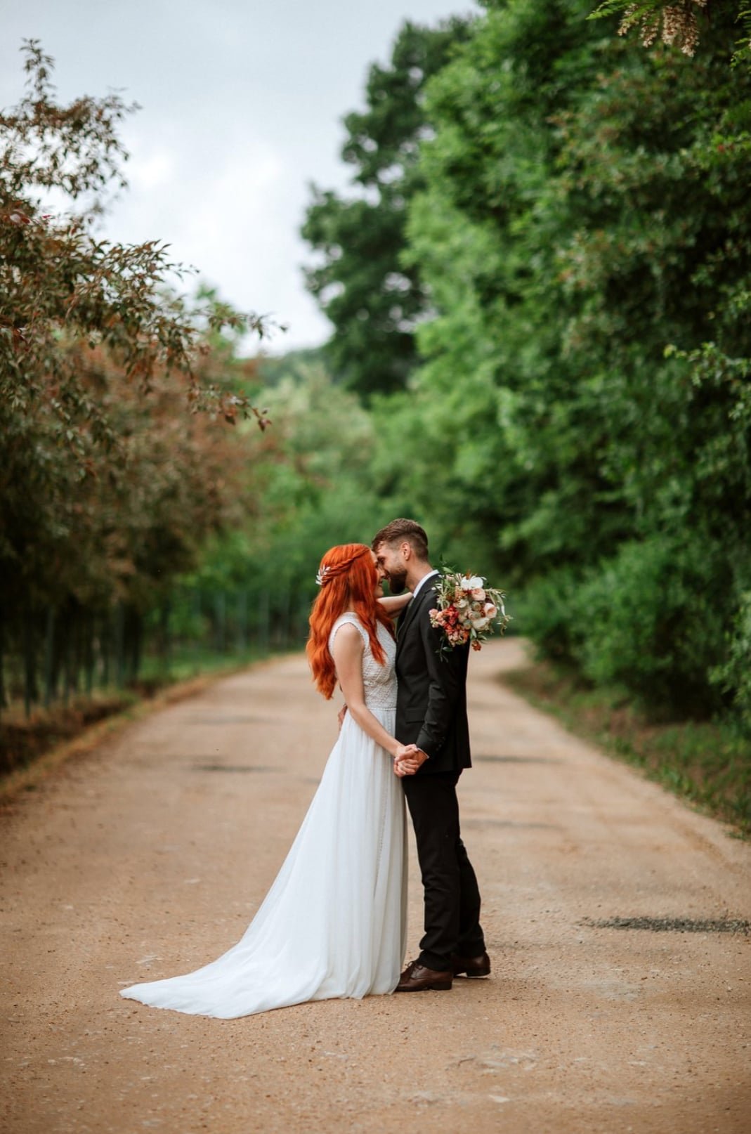 A newlywed couple standing on a dirt road surrounded by lush green trees, holding hands and about to kiss. The bride has long red hair and is wearing a white wedding dress, while the groom is dressed in a black suit. The bride is holding a bouquet of