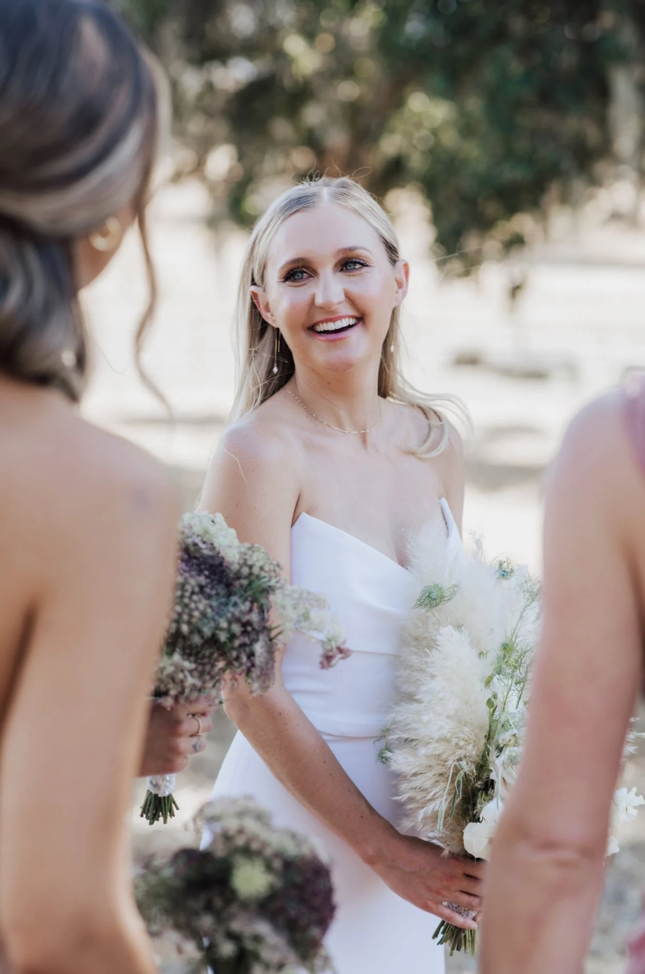 A bride with blonde hair and a white dress standing outdoors, smiling, holding a bouquet of flowers, with two women in the foreground. The background features trees with blurred foliage.