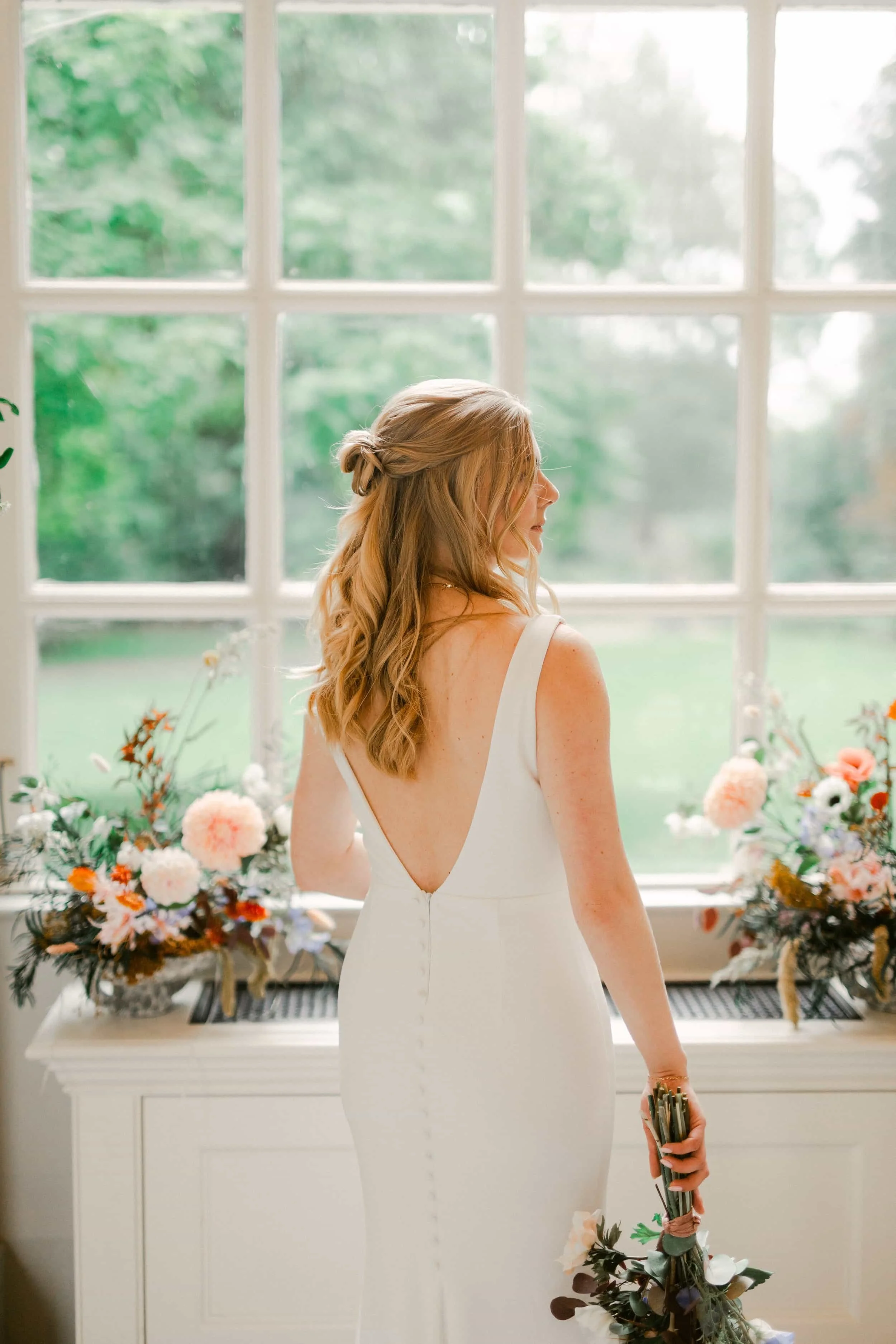 A woman with long, wavy blonde hair in a low braid, wearing a white dress with a deep open back, standing inside in front of a large window with a view of trees. She is holding a bouquet of flowers in her right hand and looking to the side. There are